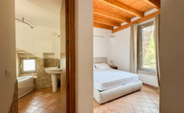 Spacious interior view of a modern bedroom with a double bed and natural light from a large window, adjacent to a stylish bathroom featuring a bathtub and sink, showcasing terracotta flooring and wooden ceiling beams.