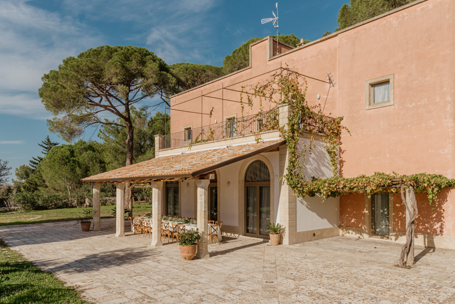 Charming Italian villa with a terracotta exterior, featuring a spacious outdoor patio with dining area, surrounded by lush greenery and tall pine trees under a clear blue sky. | Only on SopranoVillas