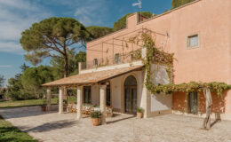Charming Italian villa with a terracotta exterior, featuring a spacious outdoor patio with dining area, surrounded by lush greenery and tall pine trees under a clear blue sky.