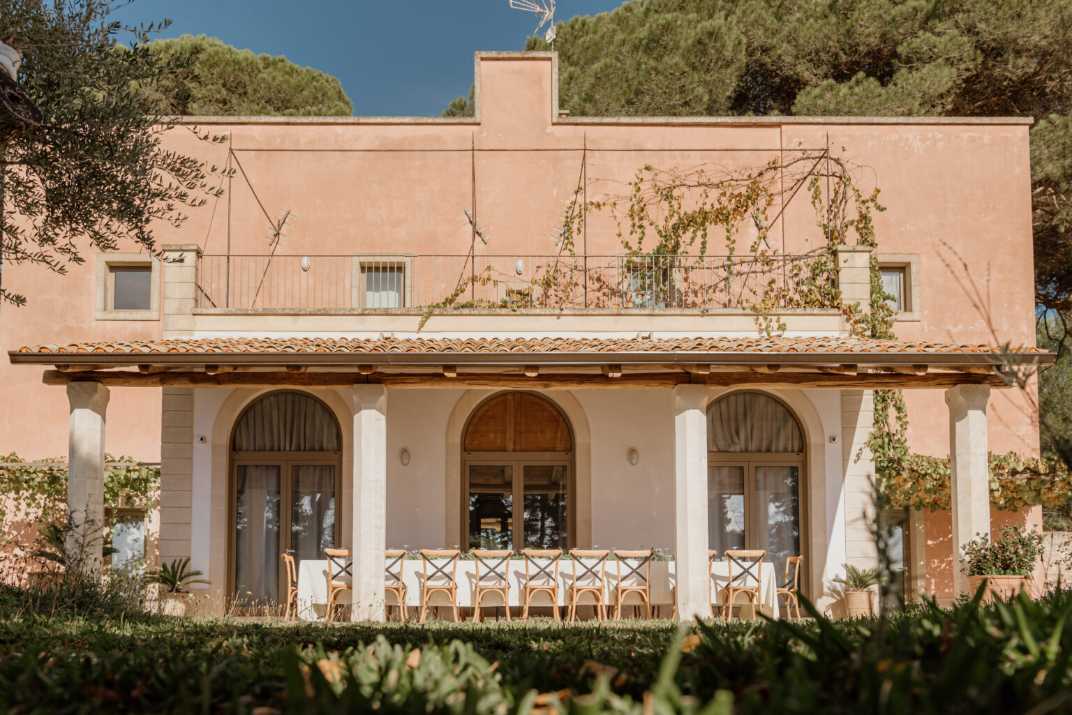 A charming pink villa featuring a spacious outdoor dining area with a long table and wooden chairs, surrounded by lush greenery and climbing vines. The villa's architecture includes arched windows and a tiled roof, set against a clear blue sky. | Only on SopranoVillas
