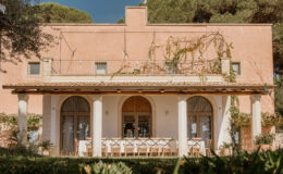 A charming pink villa featuring a spacious outdoor dining area with a long table and wooden chairs, surrounded by lush greenery and climbing vines. The villa's architecture includes arched windows and a tiled roof, set against a clear blue sky.