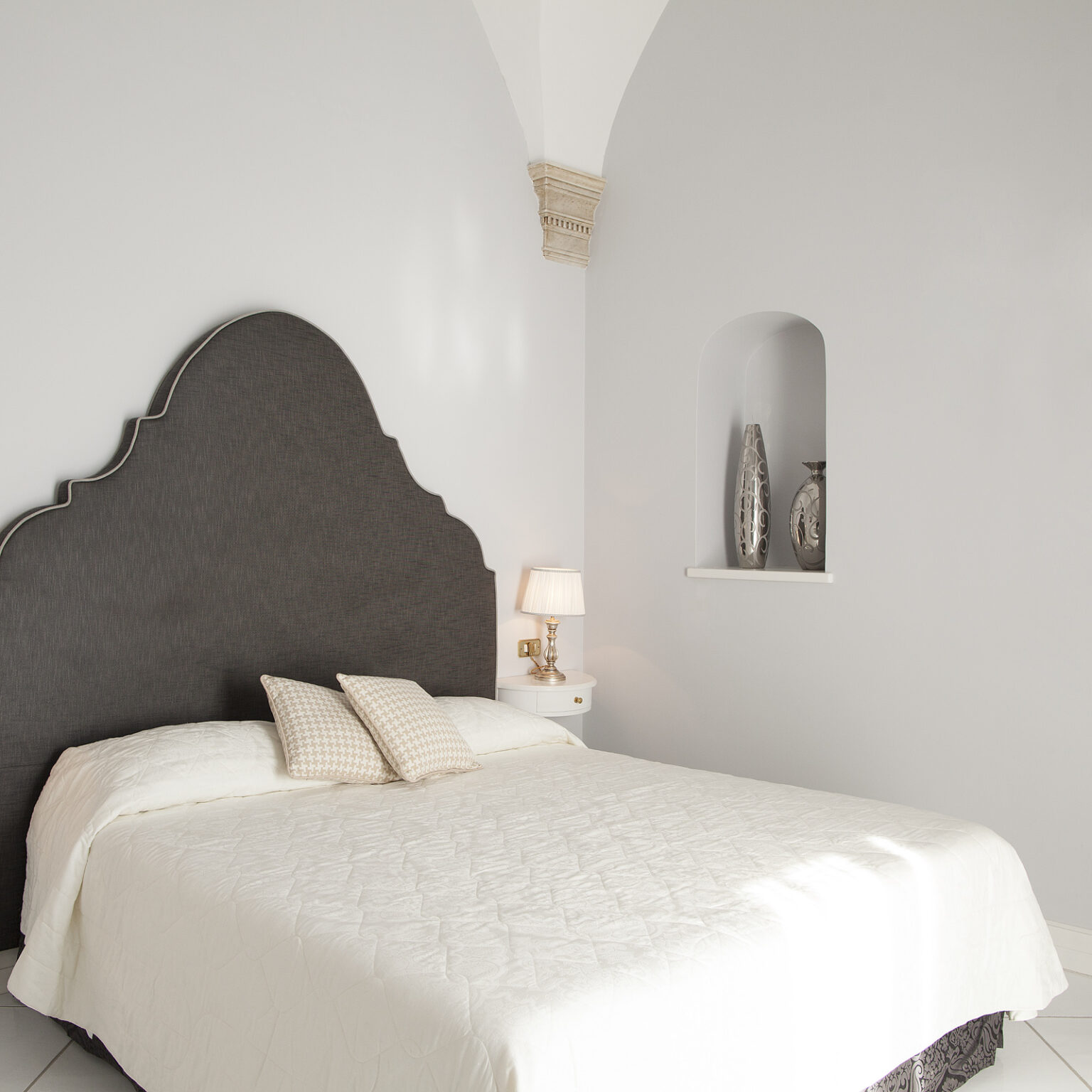 Modern bedroom interior featuring a plush bed with a decorative headboard, soft white bedding, and elegant pillows. A stylish bedside table with a lamp complements the minimalist design, while a niche showcases decorative vases against a light gray wall. Perfect for showcasing contemporary decor and relaxation. | Only on SopranoVillas