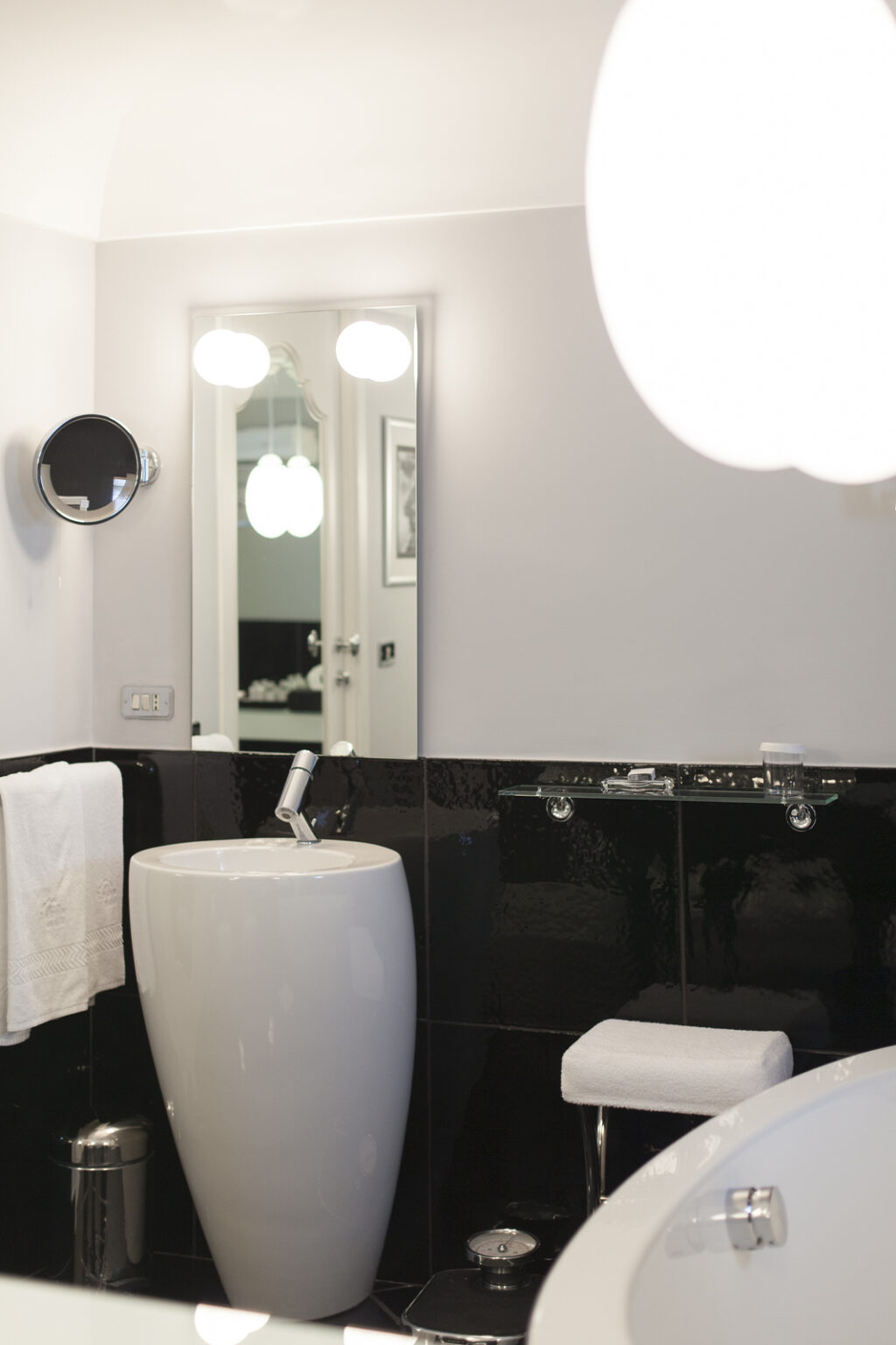 Modern bathroom featuring a sleek white pedestal sink, a round mirror, and stylish black tiles. Soft lighting illuminates the space, enhancing the contemporary design. A white towel and a small stool add functionality to the elegant decor. | Only on SopranoVillas