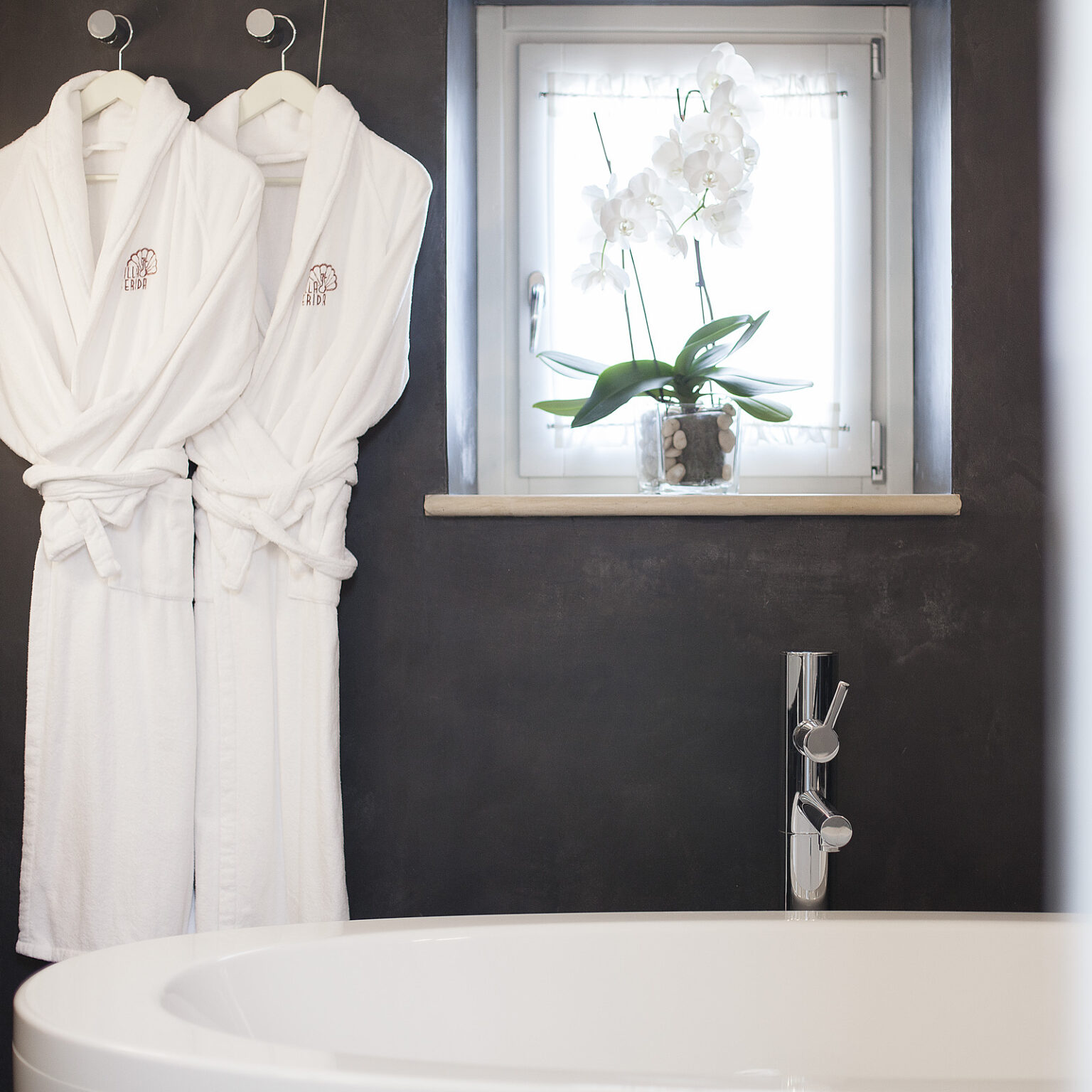 Luxurious bathroom featuring two plush white robes hanging on hooks, a modern bathtub, and a window adorned with an elegant orchid plant in a decorative pot. The dark wall contrasts beautifully with the light elements, creating a serene and inviting atmosphere. | Only on SopranoVillas