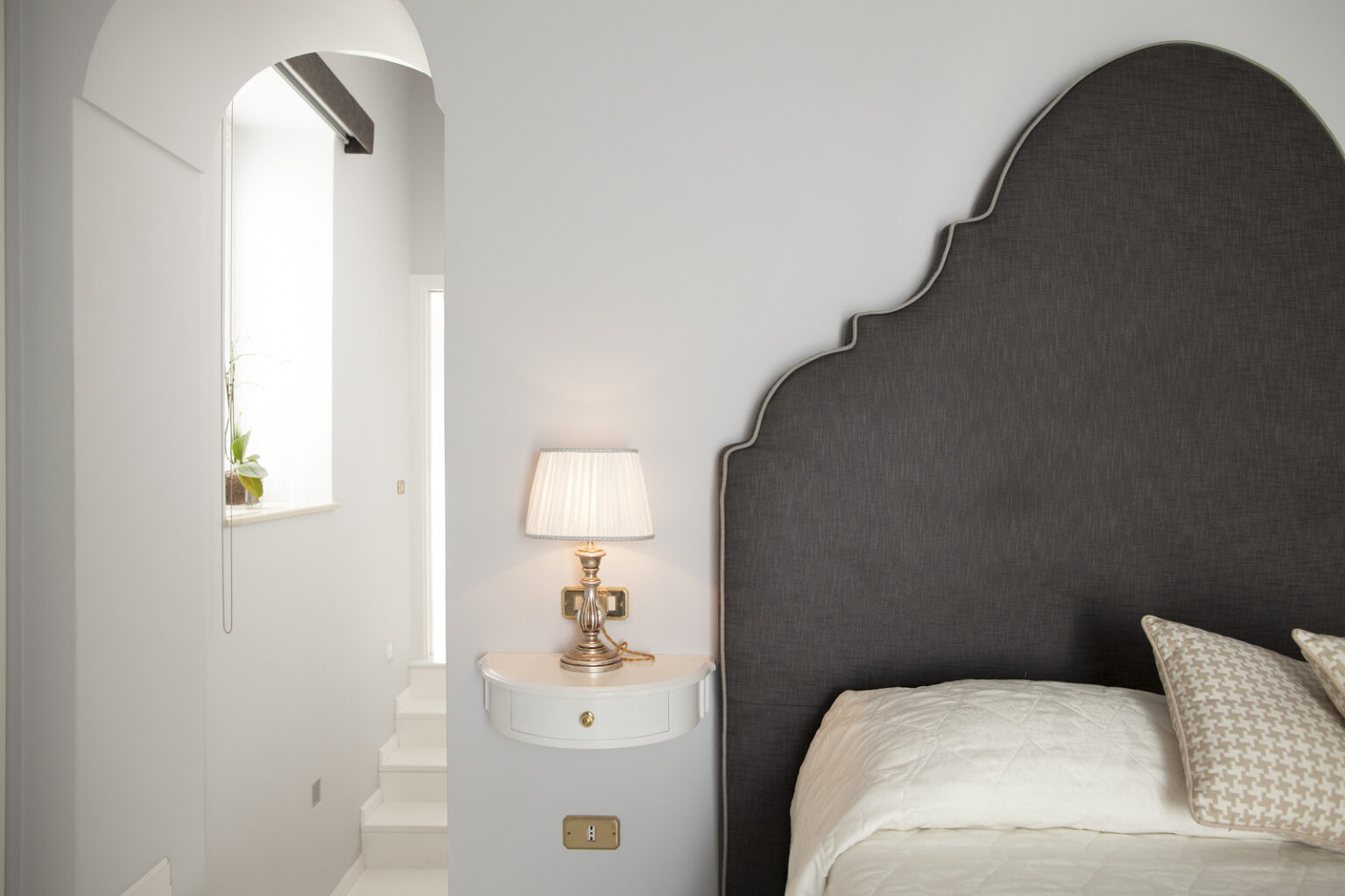 Modern bedroom interior featuring a stylish gray upholstered headboard, elegant bedside lamp, and soft bedding. The light color scheme is complemented by natural light streaming through a nearby window, enhancing the serene atmosphere. | Only on SopranoVillas