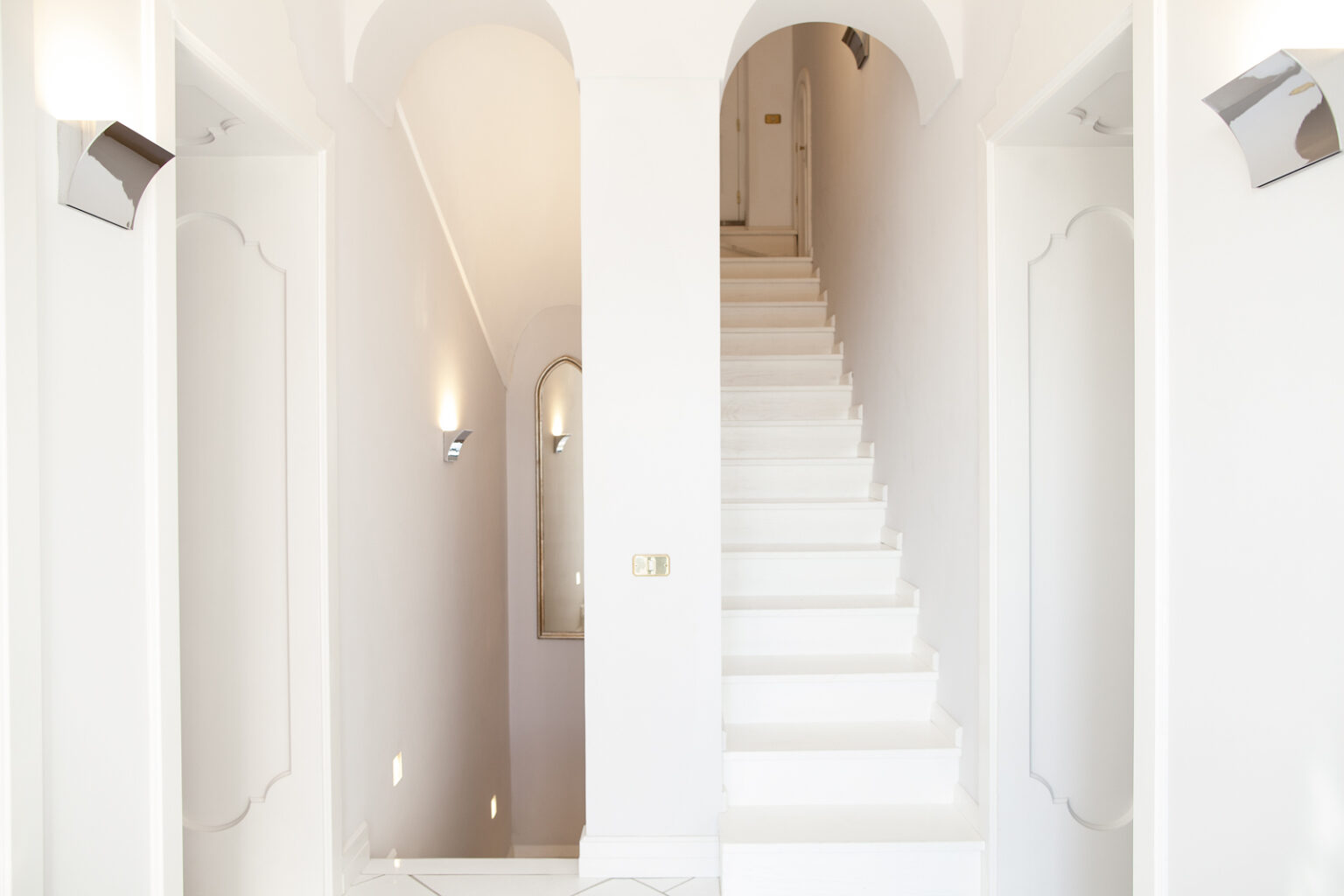 Bright and modern hallway featuring white walls, elegant archways, and a staircase leading to the upper floor, illuminated by stylish wall sconces. A mirrored accent adds depth to the space. | Only on SopranoVillas
