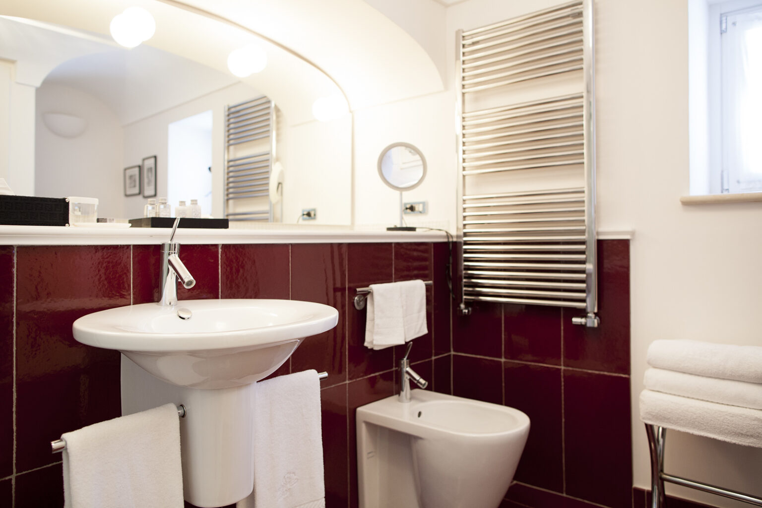 Modern bathroom featuring a white pedestal sink, bidet, and chrome towel rack. The walls are adorned with deep red tiles, and there is a large mirror above the sink, complemented by soft lighting. Fresh white towels are neatly arranged, enhancing the clean and stylish aesthetic. | Only on SopranoVillas
