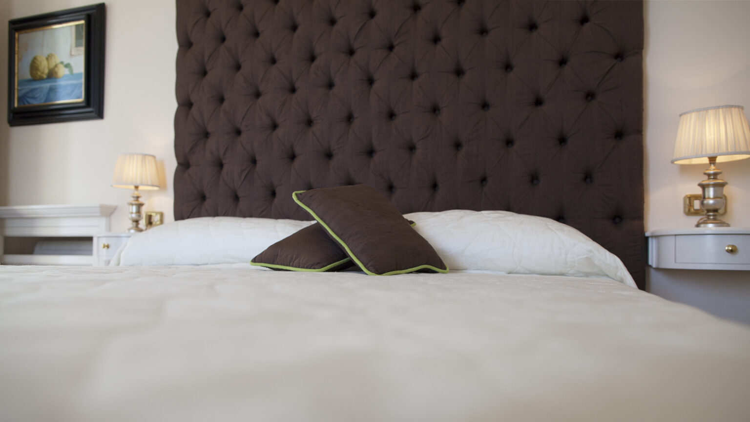 Cozy bedroom featuring a tufted brown headboard, two decorative pillows with green trim, and soft white bedding, accompanied by stylish bedside lamps and a framed artwork on the wall. Ideal for relaxation and comfort. | Only on SopranoVillas