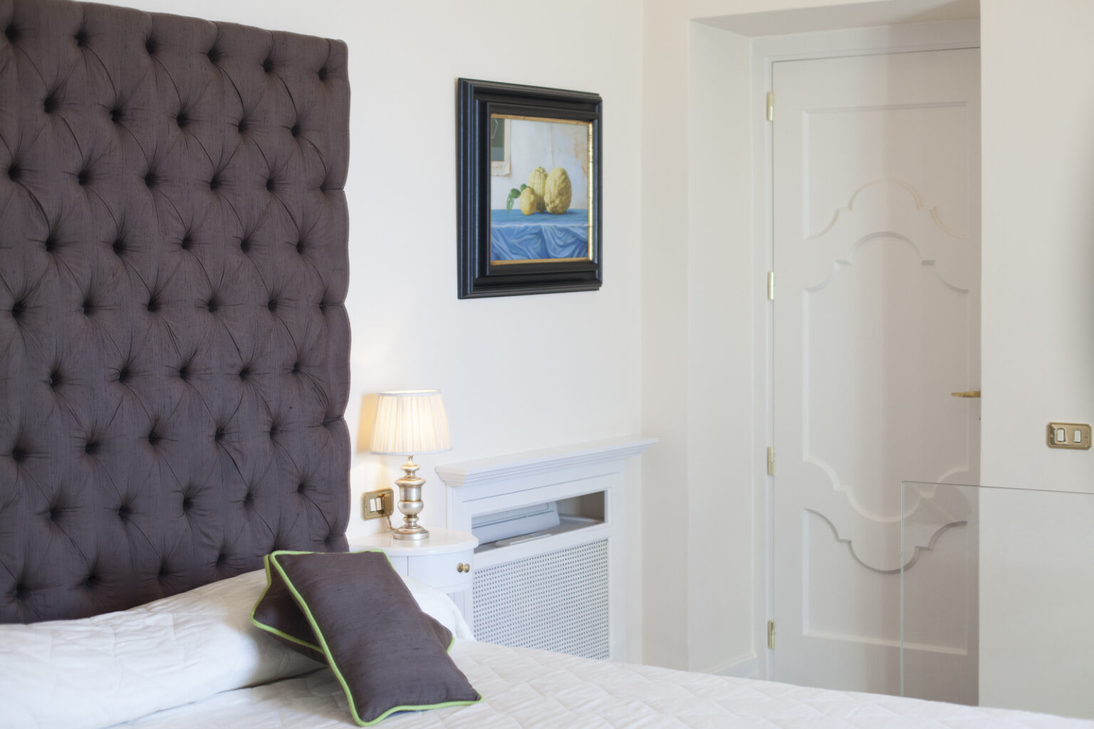Elegant bedroom interior featuring a tufted dark purple headboard, a bedside lamp, and a decorative painting. The room has a minimalist design with white walls and a stylish door, creating a serene atmosphere. | Only on SopranoVillas