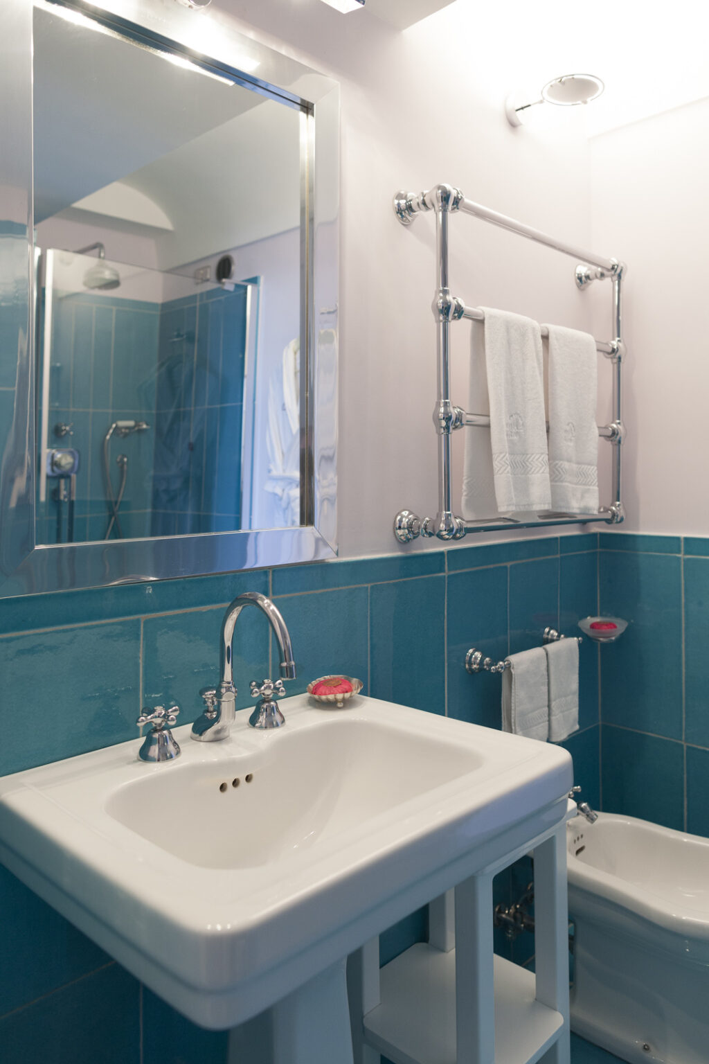 Modern bathroom featuring a white sink with chrome fixtures, a bidet, and blue tiled walls. The space includes a large mirror, towel rack, and decorative soap, creating a clean and stylish look. | Only on SopranoVillas