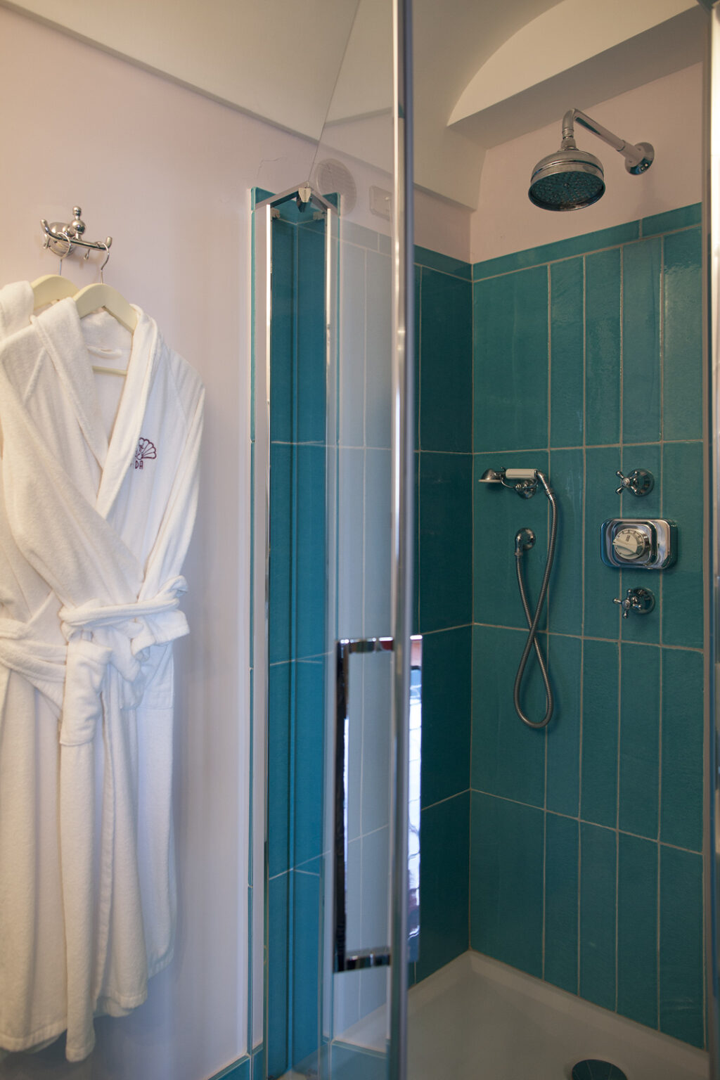 Modern shower with turquoise tiles, chrome fixtures, and a glass enclosure, featuring a white bathrobe hanging nearby, creating a stylish and inviting bathroom atmosphere. | Only on SopranoVillas