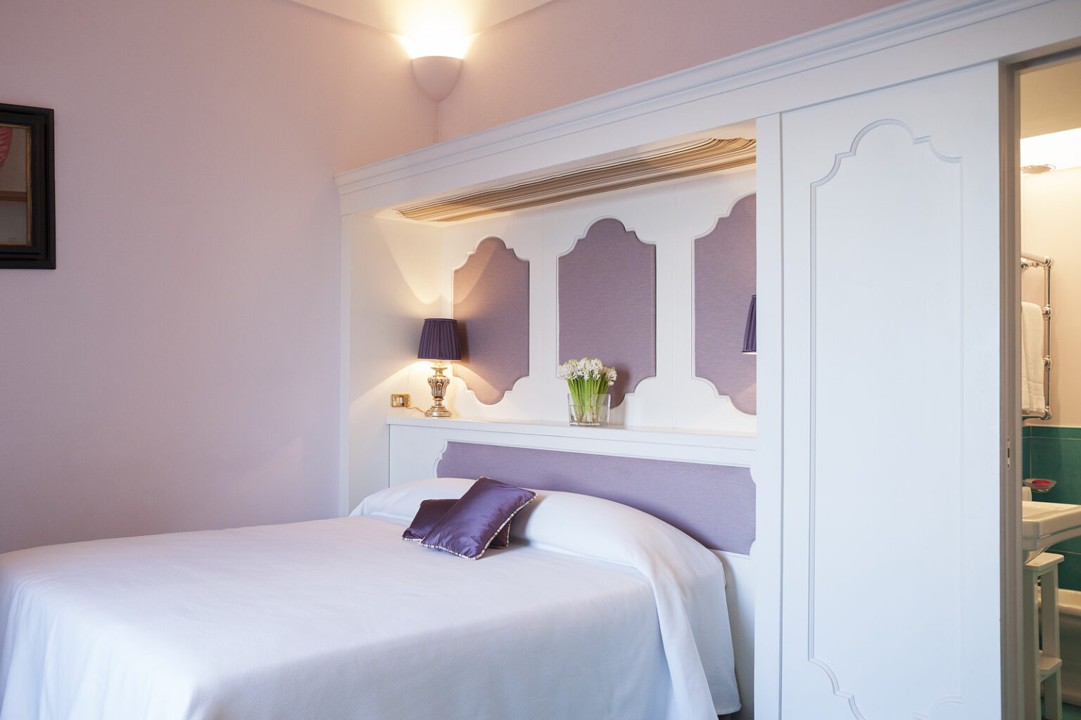 Cozy bedroom featuring a white bed with a purple accent pillow, soft pink walls, and a stylish headboard design. The room is illuminated by a wall-mounted light and includes a small vase of flowers on a shelf, with a glimpse of a bathroom visible in the background. | Only on SopranoVillas