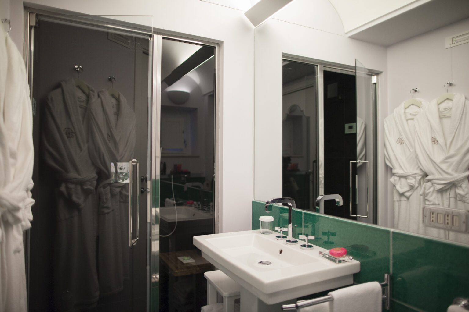 Modern bathroom interior featuring a sleek shower enclosure, stylish sink with multiple faucets, and plush bathrobes hanging nearby. The vibrant green tiles add a pop of color, enhancing the contemporary design. | Only on SopranoVillas