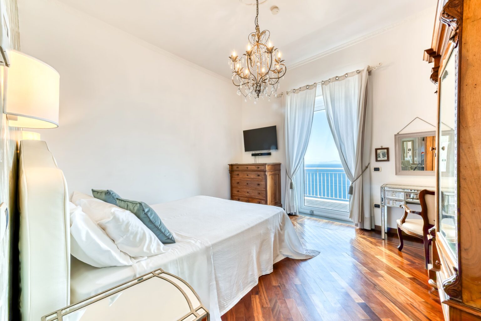 Bright and airy bedroom featuring a chandelier, hardwood floors, a large window with sheer curtains, a flat-screen TV, and elegant furniture, creating a cozy and inviting atmosphere. | Only on SopranoVillas