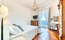 Bright and airy bedroom featuring a chandelier, hardwood floors, a large window with sheer curtains, a flat-screen TV, and elegant furniture, creating a cozy and inviting atmosphere.