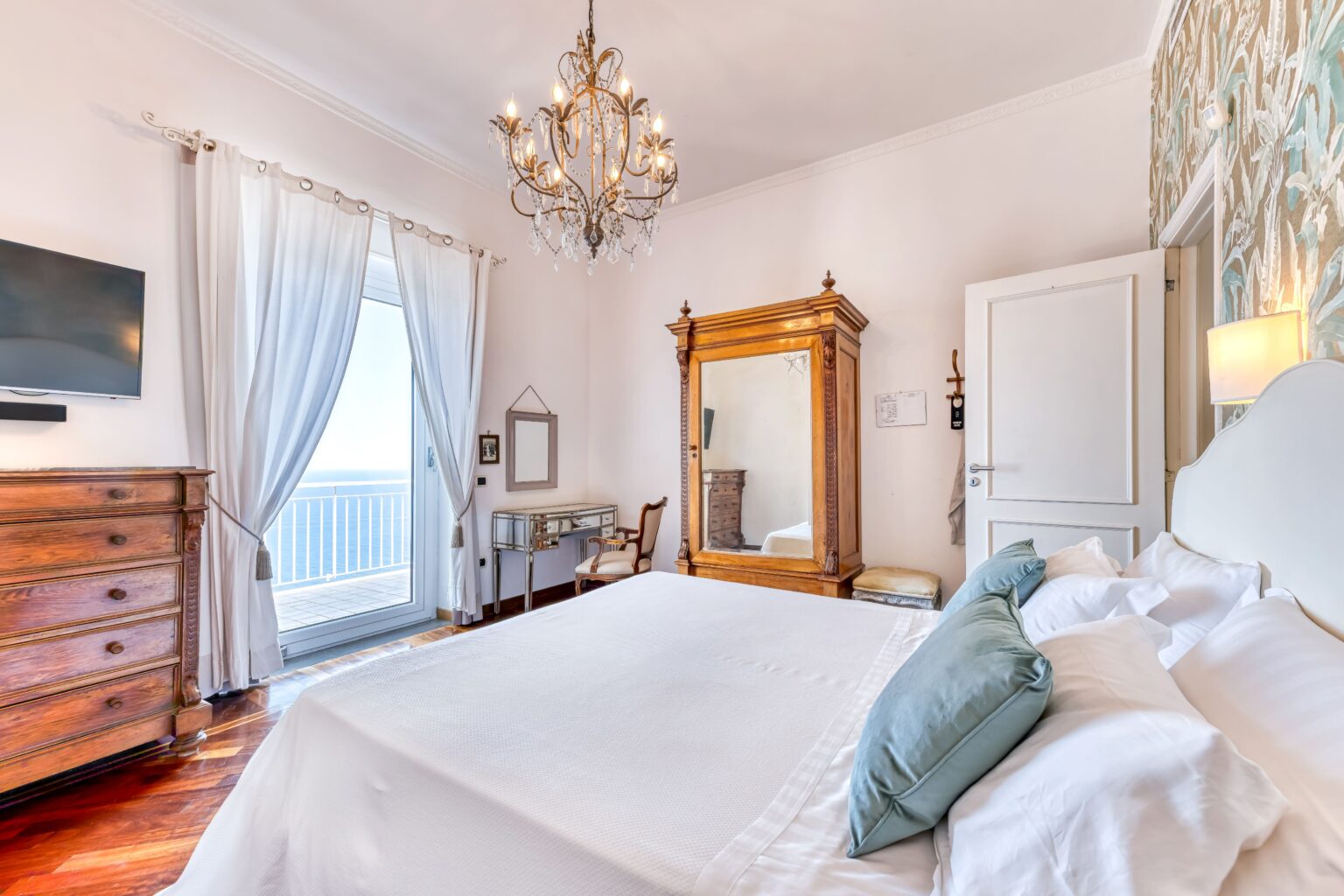 Elegant bedroom featuring a king-size bed with decorative pillows, a vintage wooden dresser, a large mirror, and a chandelier. Natural light floods the room through sheer curtains, highlighting the stylish decor and inviting atmosphere. | Only on SopranoVillas