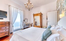 Elegant bedroom featuring a king-size bed with decorative pillows, a vintage wooden dresser, a large mirror, and a chandelier. Natural light floods the room through sheer curtains, highlighting the stylish decor and inviting atmosphere.