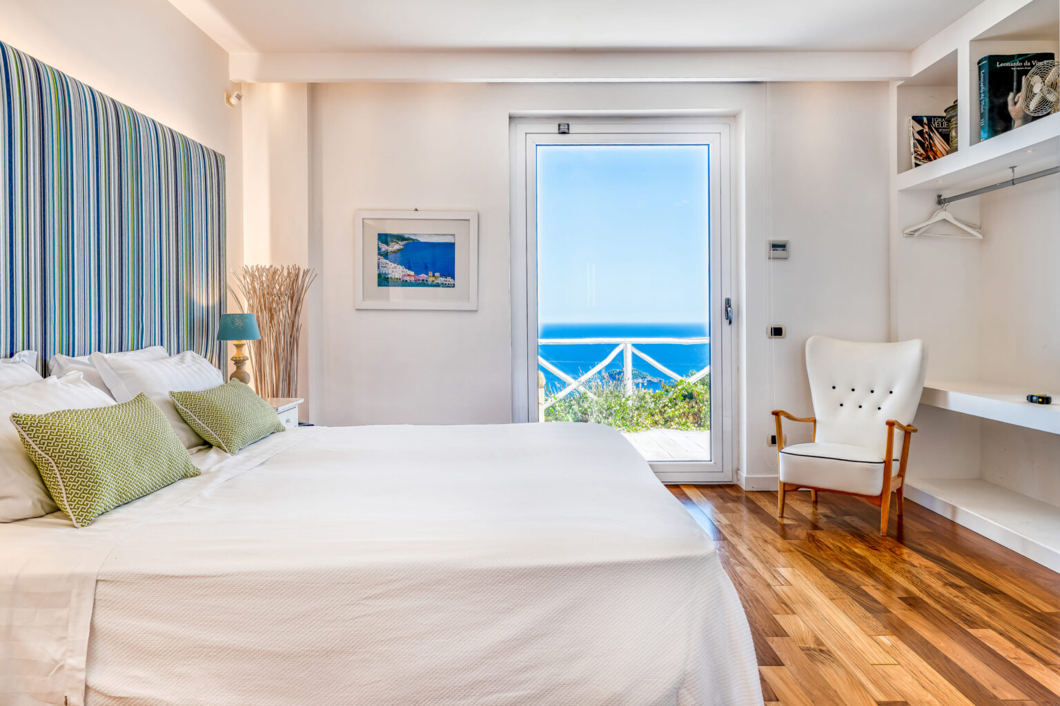 Modern coastal bedroom with a large bed, green accent pillows, and a chair, featuring a view of the ocean through a sliding glass door. The room has natural light, wooden flooring, and stylish decor elements. Ideal for beach vacation rentals. | Only on SopranoVillas