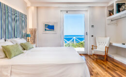Modern coastal bedroom with a large bed, green accent pillows, and a chair, featuring a view of the ocean through a sliding glass door. The room has natural light, wooden flooring, and stylish decor elements. Ideal for beach vacation rentals.
