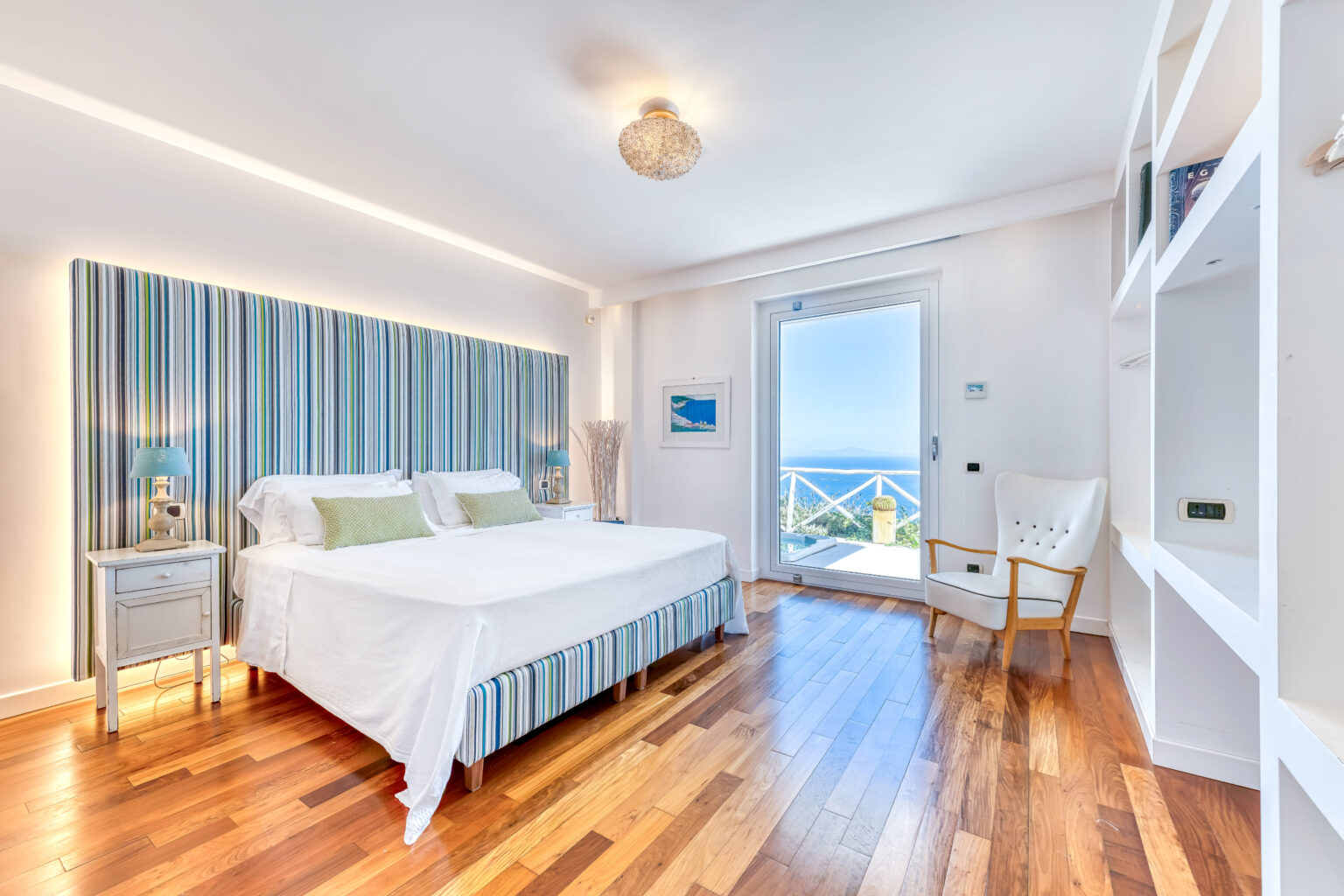 Modern bedroom featuring a striped headboard, white bedding, and hardwood flooring, with a scenic ocean view from the window and stylish decor elements. | Only on SopranoVillas