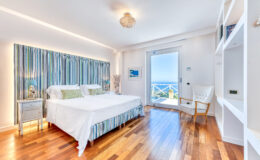 Modern bedroom featuring a striped headboard, white bedding, and hardwood flooring, with a scenic ocean view from the window and stylish decor elements.