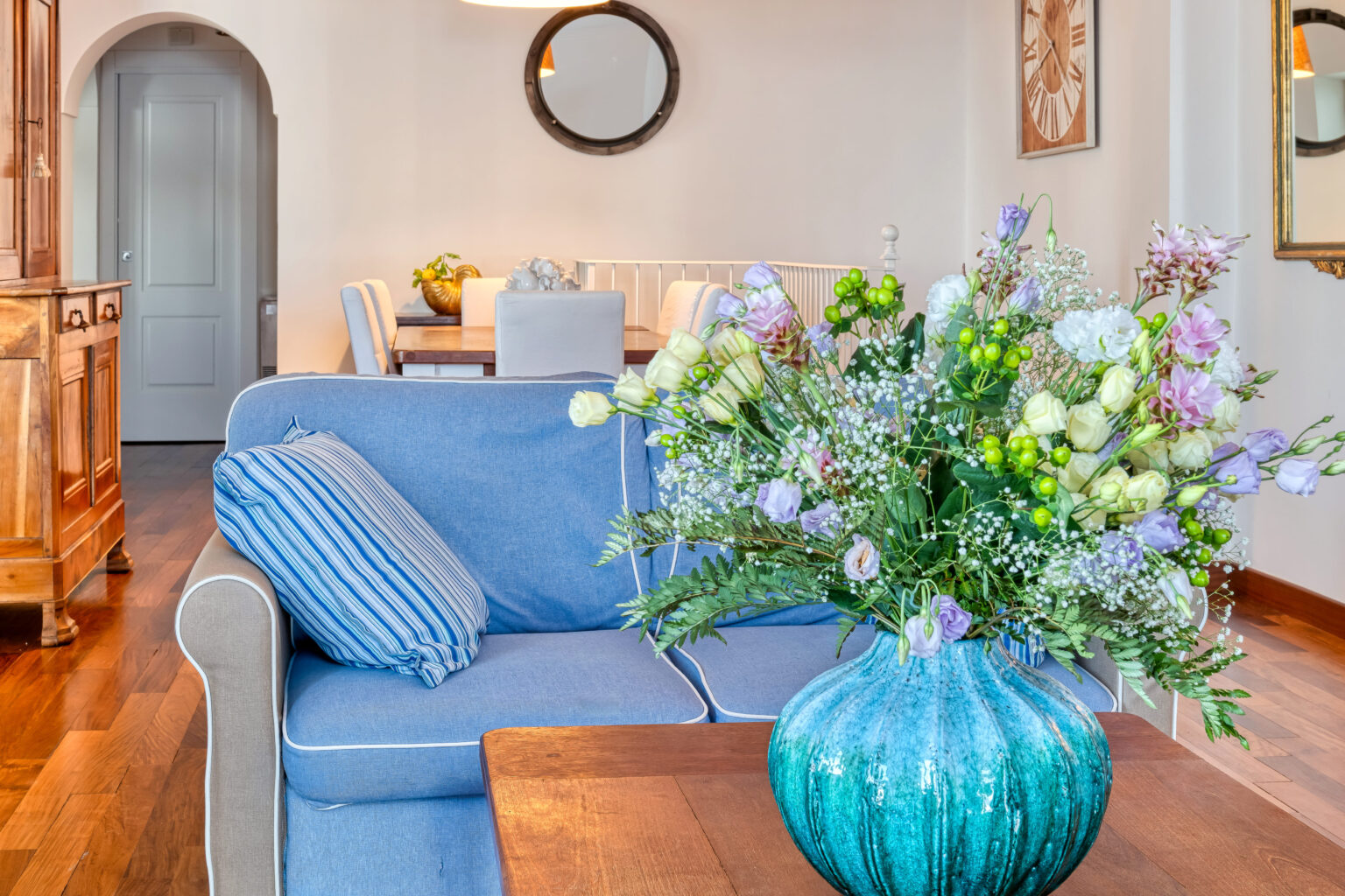 Cozy living room featuring a blue striped couch adorned with decorative pillows, a vibrant turquoise vase filled with fresh flowers, and a wooden coffee table, creating a warm and inviting atmosphere. | Only on SopranoVillas