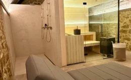 Modern spa interior featuring a glass sauna, stone walls, and a relaxing lounge area with a gray chaise lounge. The design combines natural materials and soft lighting for a tranquil atmosphere.