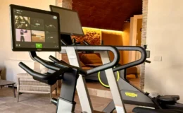 Modern Technogym exercise bike with interactive touchscreen display, set in a spacious and stylish gym area featuring an arched brick wall and cozy seating.