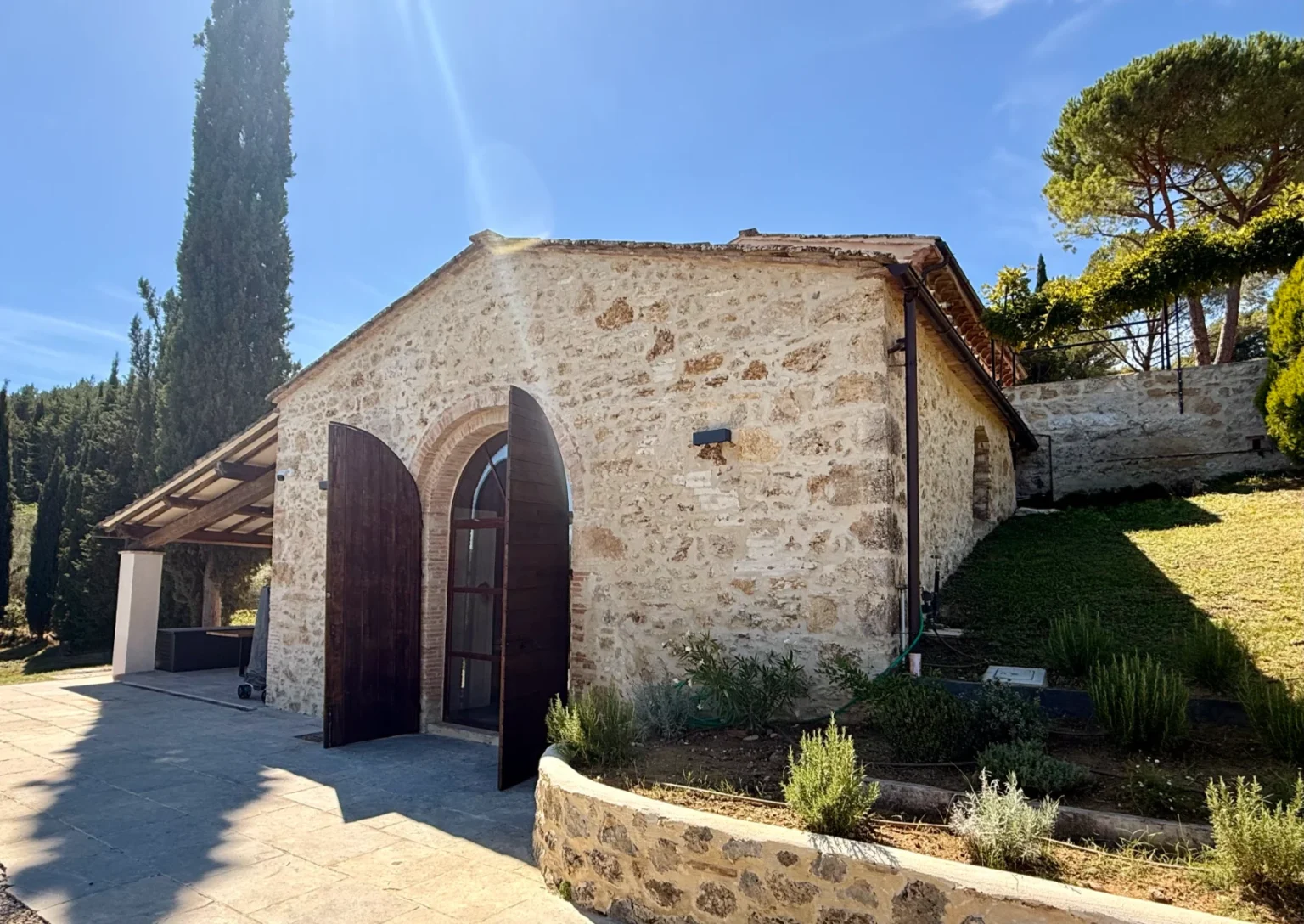 Stone cottage with wooden double doors set in a serene landscape, surrounded by greenery and tall trees under a clear blue sky. The structure features a rustic design and is complemented by a stone pathway and landscaped garden. Ideal for nature lovers and those seeking a peaceful retreat. | Only on SopranoVillas