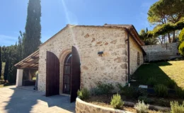 Stone cottage with wooden double doors set in a serene landscape, surrounded by greenery and tall trees under a clear blue sky. The structure features a rustic design and is complemented by a stone pathway and landscaped garden. Ideal for nature lovers and those seeking a peaceful retreat.