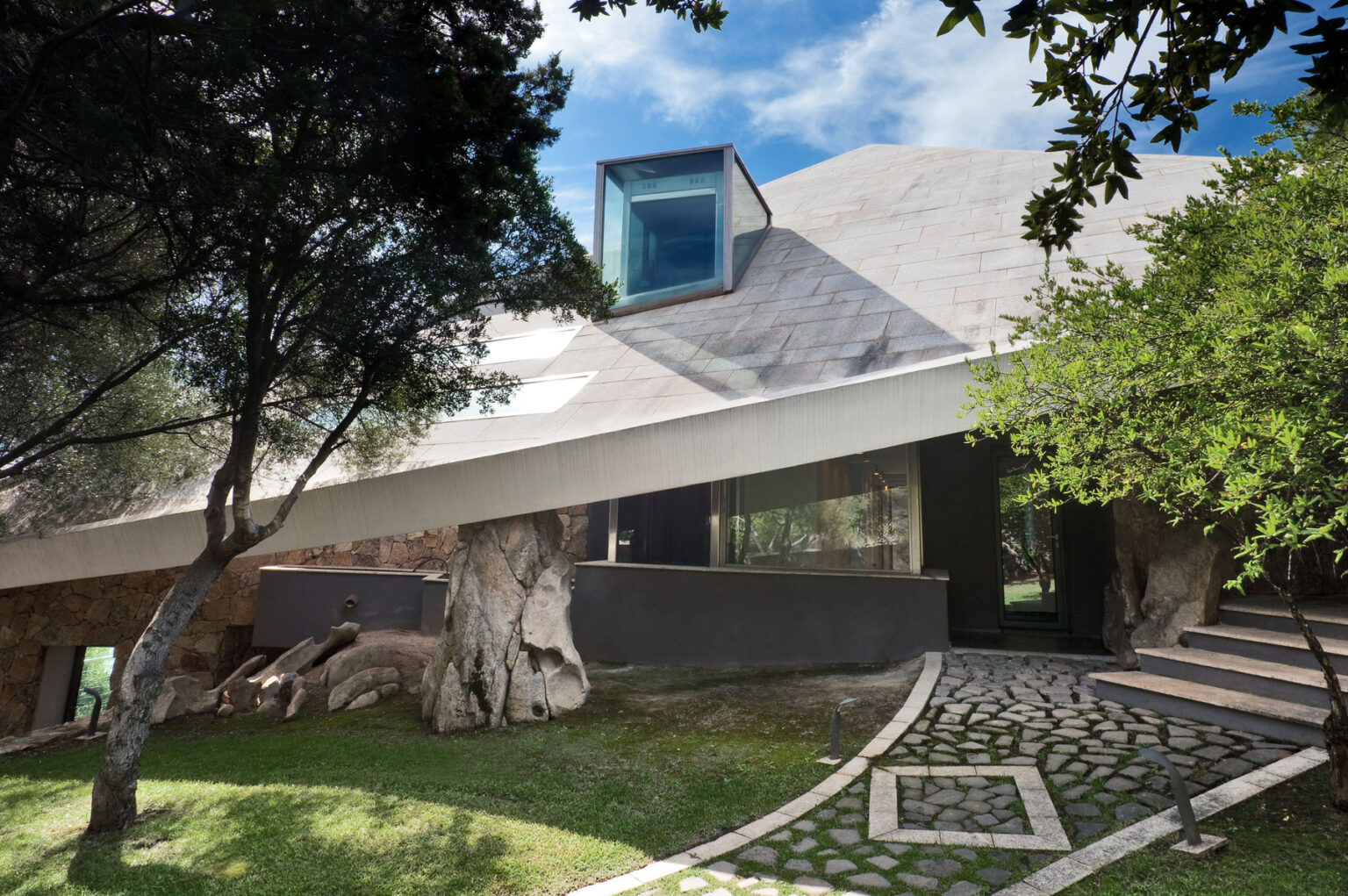 Modern architectural design of a unique house featuring a sloped roof, large glass windows, and a blend of natural elements, surrounded by greenery and stone pathways. | Only on SopranoVillas