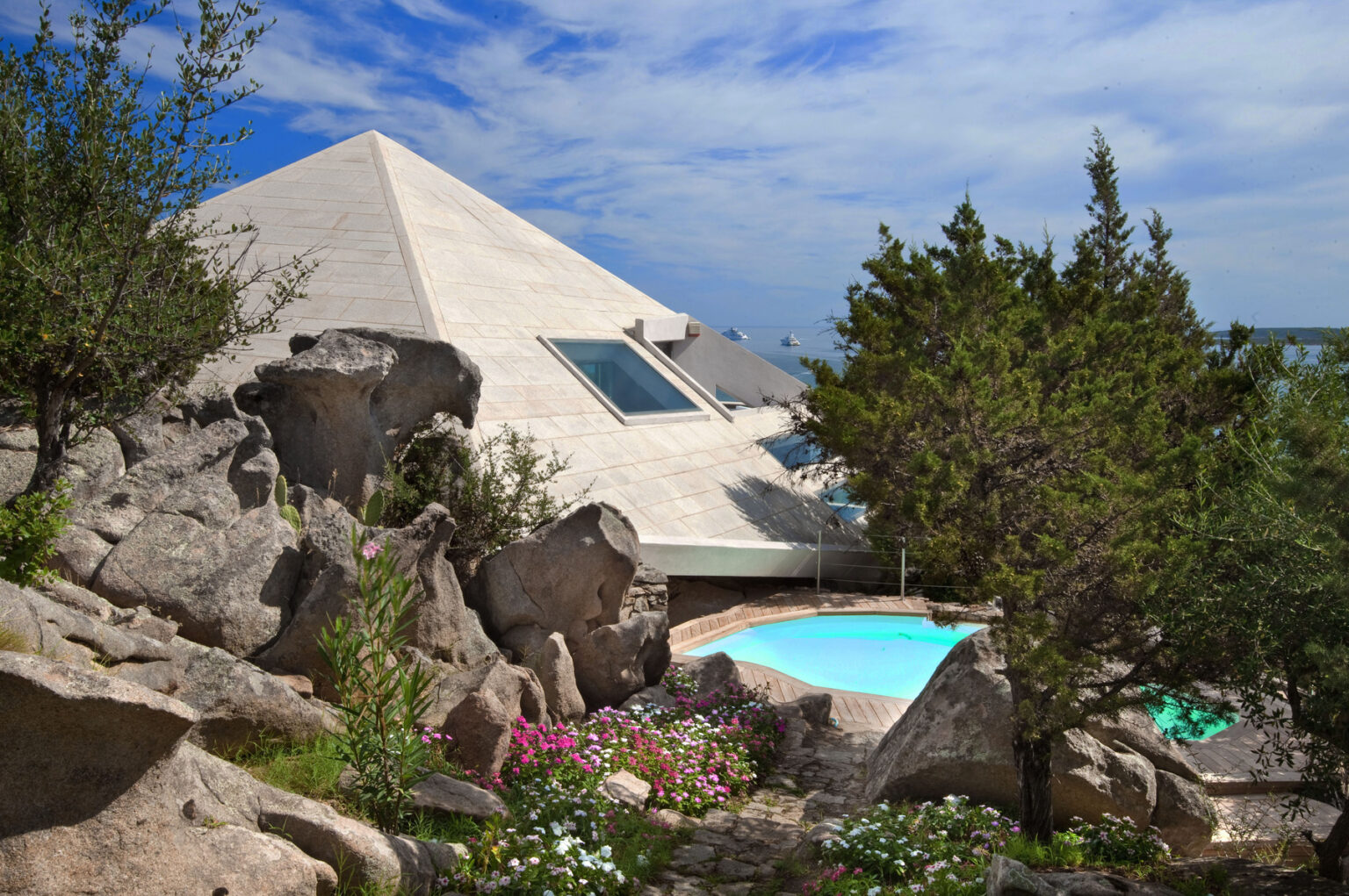 Modern pyramid-shaped house surrounded by natural rock formations and greenery, featuring a swimming pool and vibrant flowers in the foreground. | Only on SopranoVillas