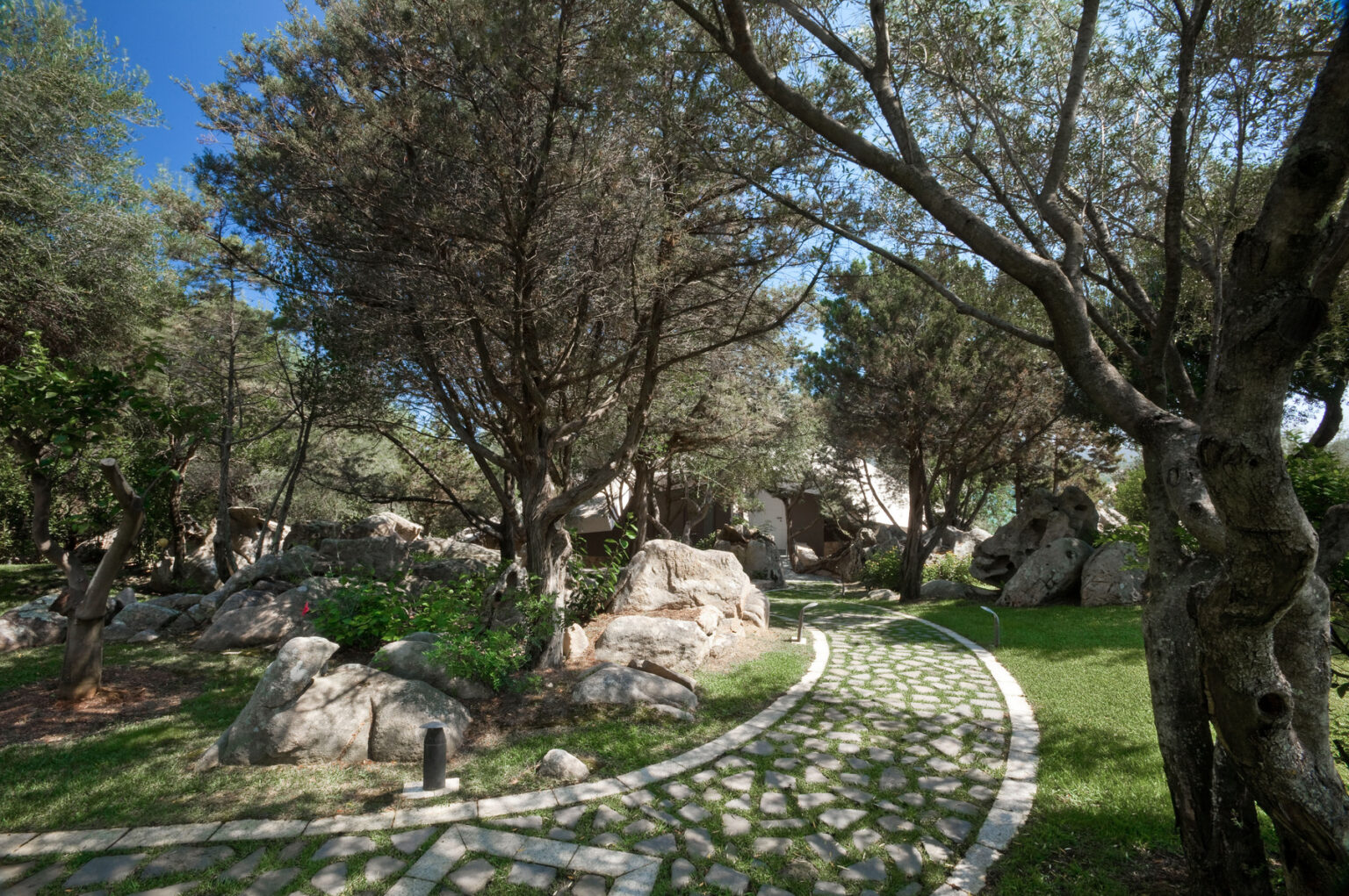 Scenic garden pathway winding through lush greenery and trees, featuring natural stone elements and a clear blue sky. | Only on SopranoVillas