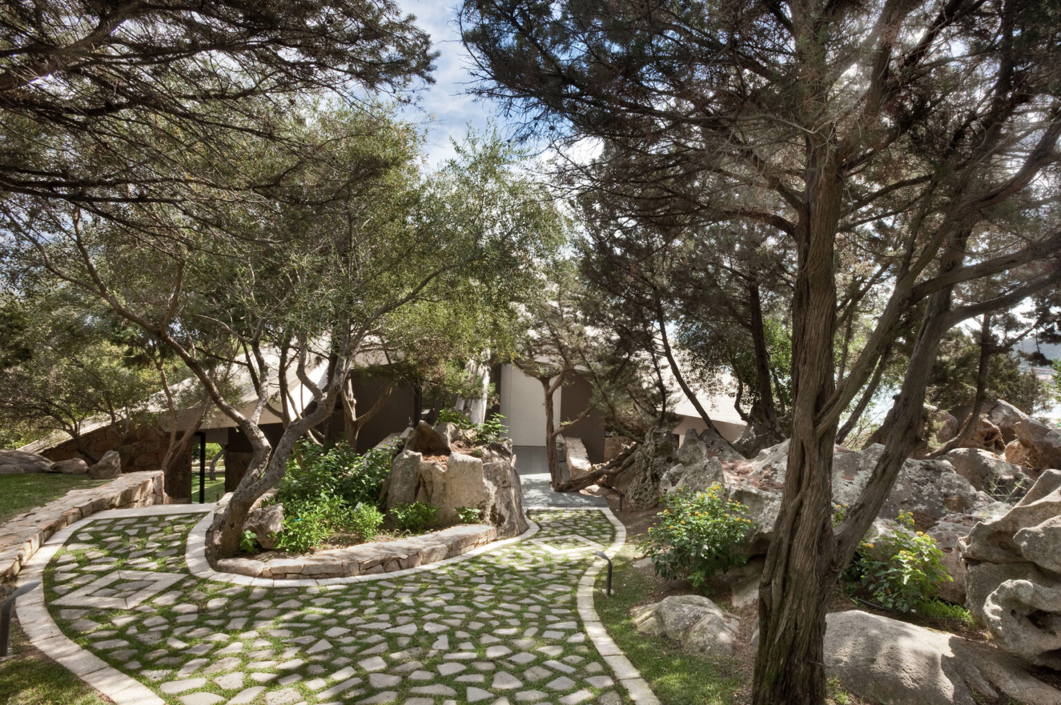 Scenic pathway leading to a modern architectural structure surrounded by lush greenery and natural rock formations, with sunlight filtering through trees. | Only on SopranoVillas