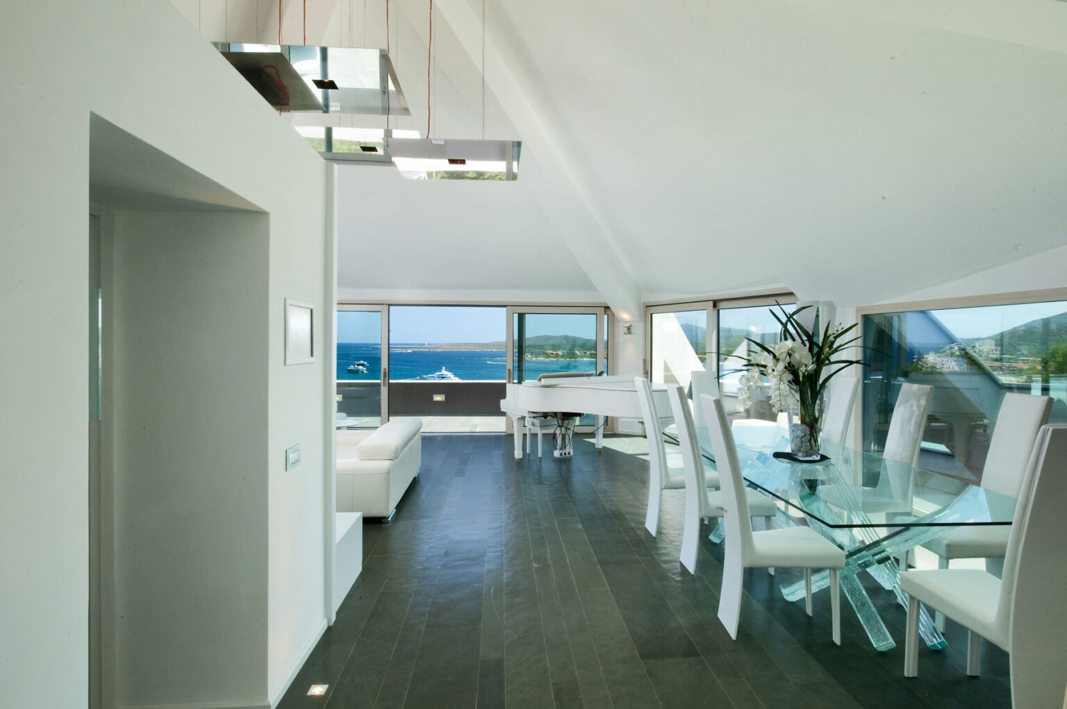Modern open-concept living space featuring a stylish dining area with a glass table and white chairs, large windows showcasing a scenic ocean view, and sleek, minimalist decor. | Only on SopranoVillas