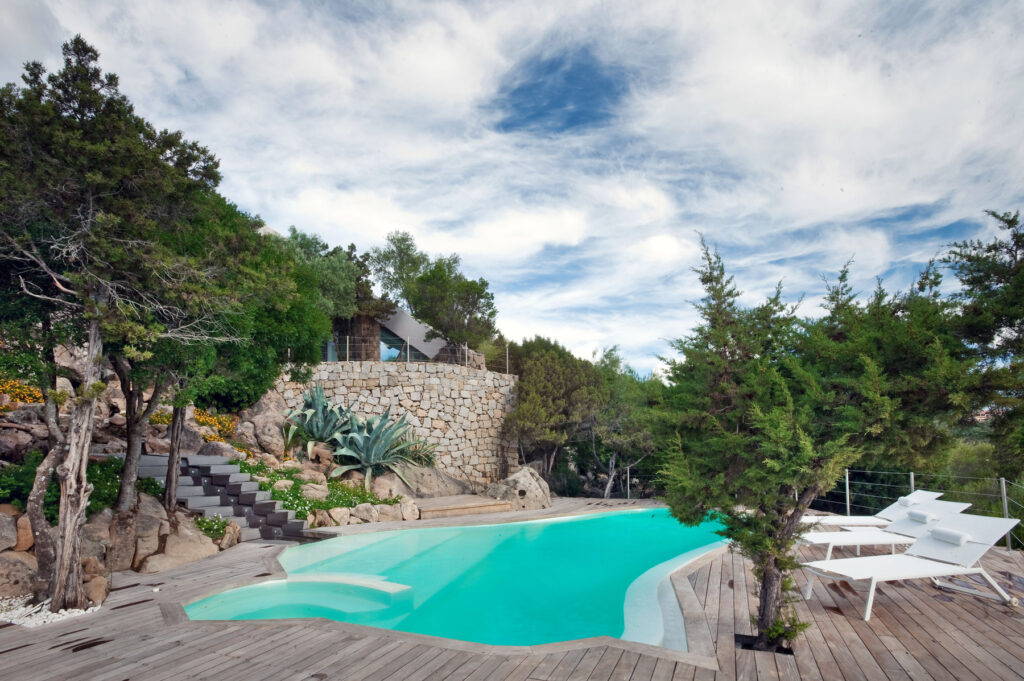 Modern outdoor swimming pool surrounded by lush greenery and natural stone landscaping, featuring wooden decking and lounge chairs, under a partly cloudy sky. Ideal for relaxation and leisure in a scenic setting. | Only on SopranoVillas