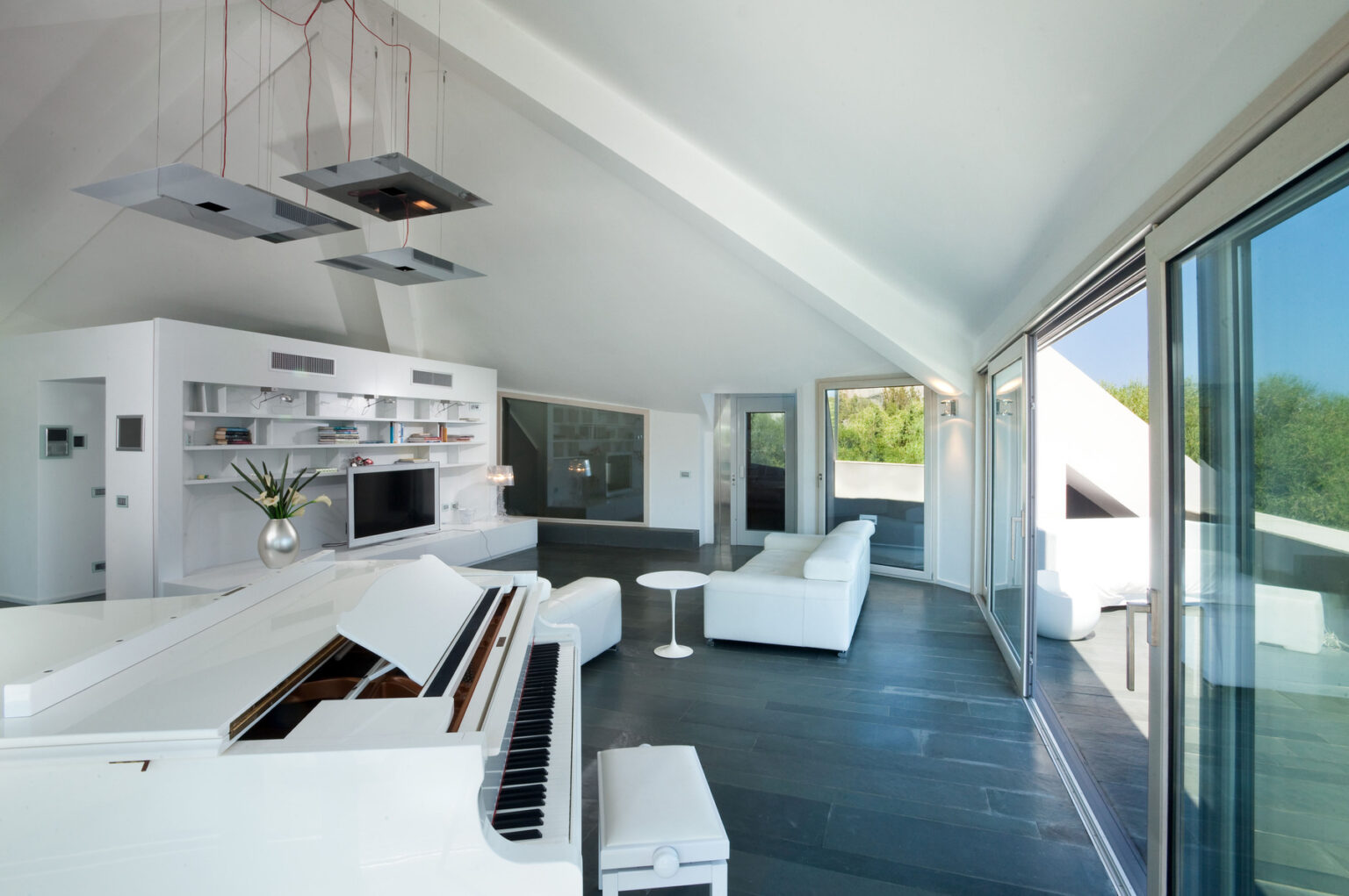 Modern living room featuring a white grand piano, sleek furniture, and large windows that offer natural light and views of the outdoors. The minimalist design includes a contemporary kitchen area with stylish lighting fixtures. | Only on SopranoVillas