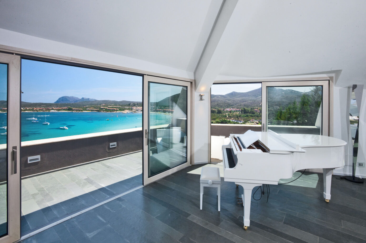 Bright and airy living space featuring a white grand piano, with large glass doors showcasing a stunning view of the turquoise sea and mountains in the background. Perfect for coastal luxury living. | Only on SopranoVillas