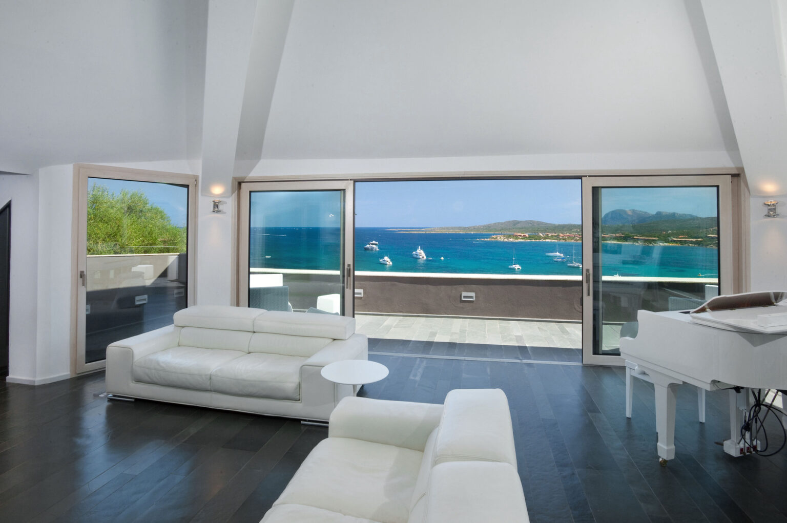 Modern living room with white leather sofas and a grand piano, featuring large windows that showcase a stunning ocean view and lush greenery. Ideal for luxury real estate listings. | Only on SopranoVillas