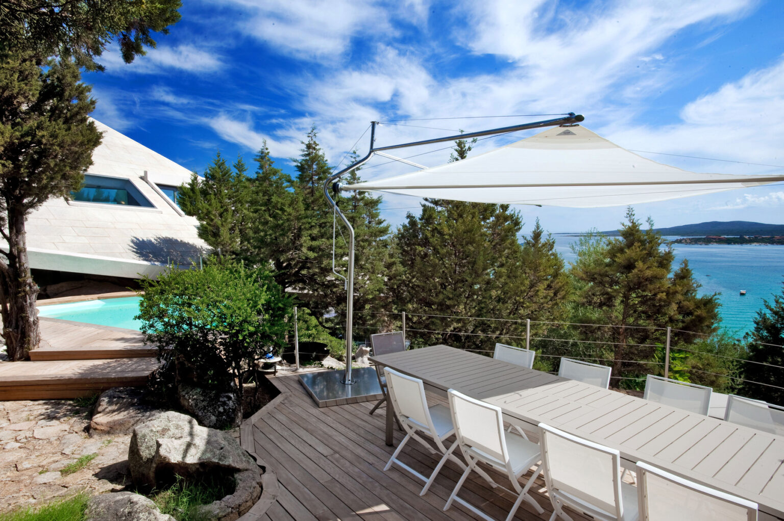 Modern outdoor dining area with a large table and white chairs, surrounded by lush greenery and overlooking a serene pool and ocean view under a bright blue sky. | Only on SopranoVillas
