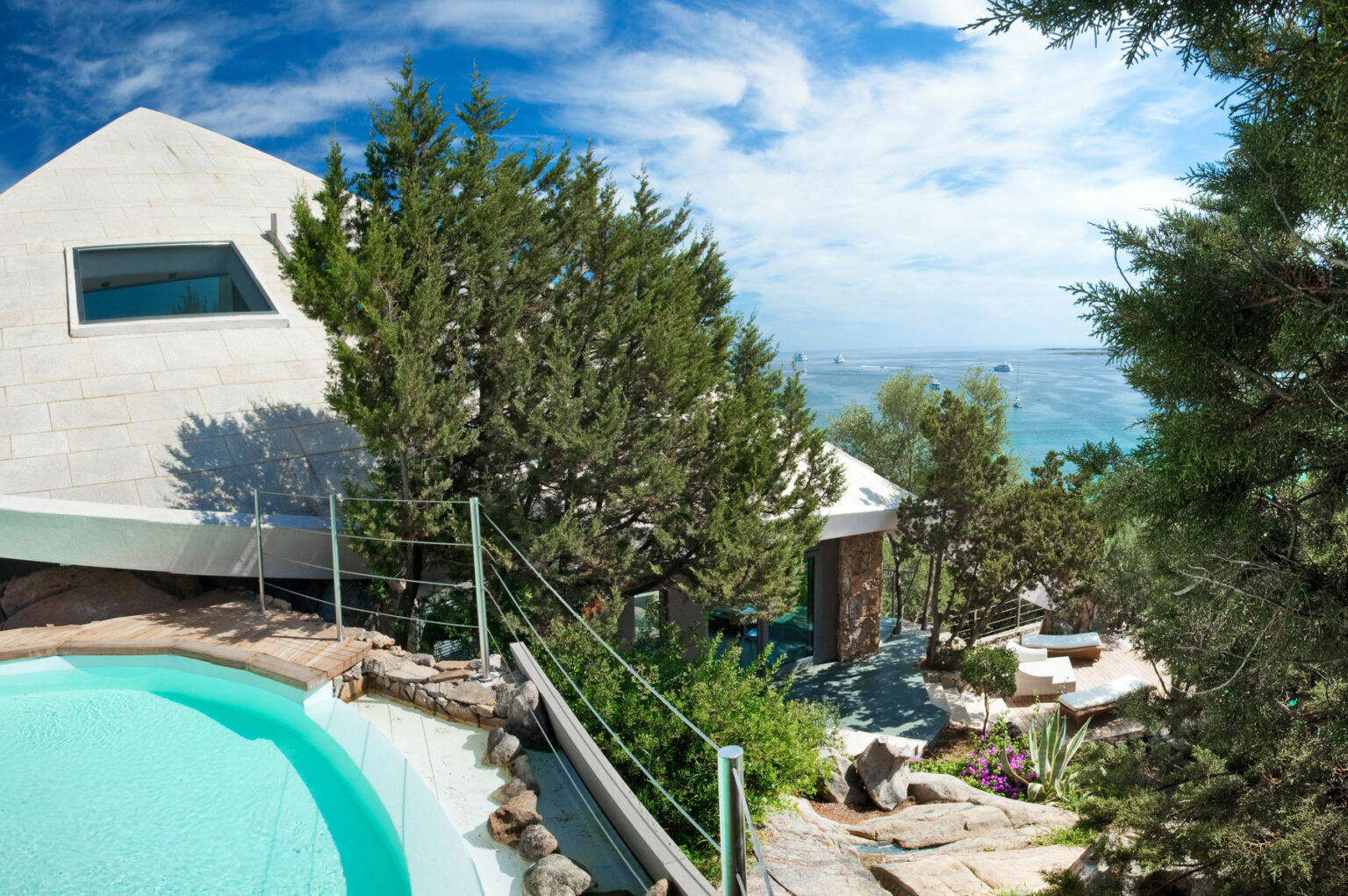 Modern architectural villa with a swimming pool, surrounded by lush greenery and scenic ocean views, featuring boats in the distance and a clear blue sky. Ideal for luxury retreats and vacation getaways. | Only on SopranoVillas