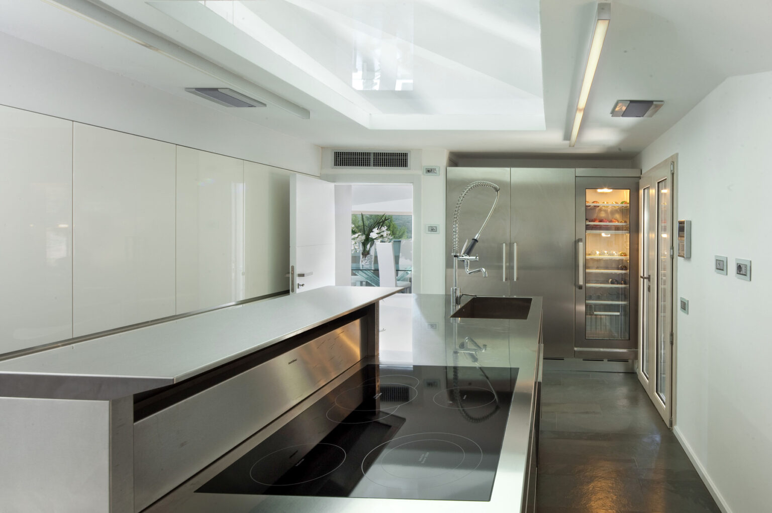 Modern kitchen interior featuring sleek white cabinetry, a central island with a stainless steel countertop, and high-end appliances, illuminated by natural light from a skylight. | Only on SopranoVillas