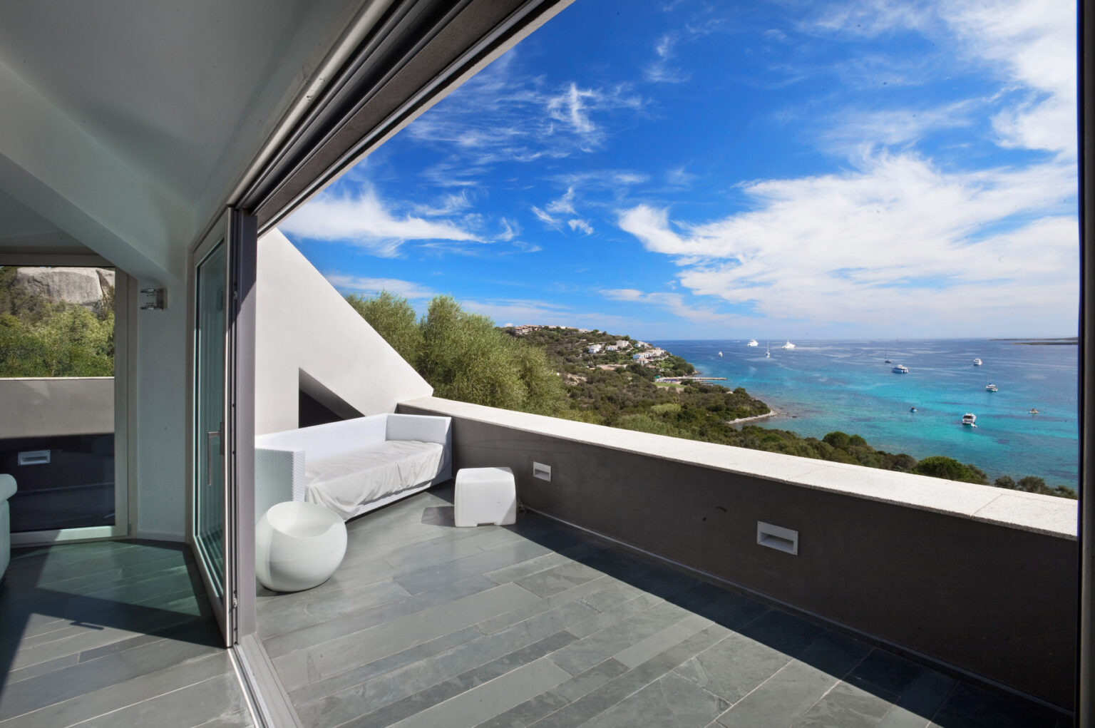 Modern balcony view overlooking a serene coastal landscape with clear blue waters and boats, showcasing a stylish outdoor seating area and vibrant sky. | Only on SopranoVillas