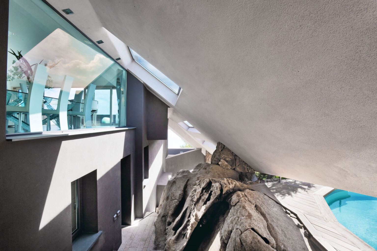 Modern architectural design showcasing a home integrated with natural rock formations, featuring large glass windows and an angled roof. The image highlights the seamless blend of indoor and outdoor spaces, including a swimming pool area. | Only on SopranoVillas