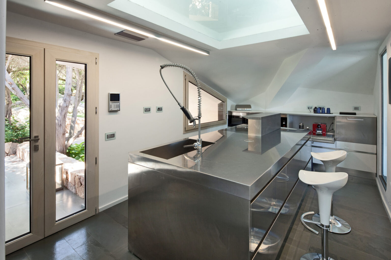 Modern kitchen interior featuring stainless steel countertops, sleek cabinetry, and a unique faucet design. Large windows provide natural light and a view of the outdoor landscape. Minimalist decor with white bar stools and a contemporary design aesthetic. | Only on SopranoVillas