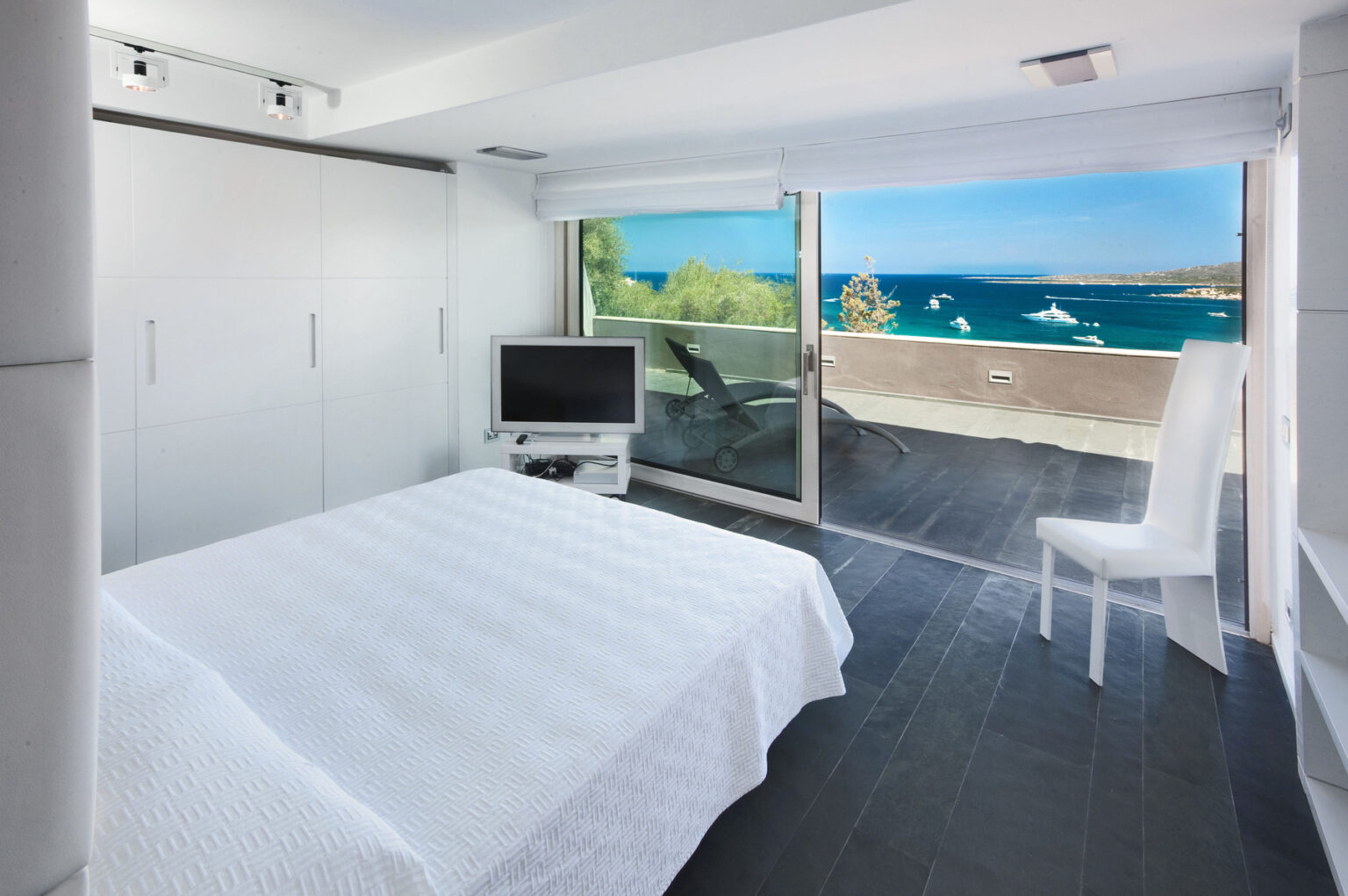 Modern bedroom with a white bedspread, flat-screen TV, and a large glass door leading to a balcony with a stunning ocean view. The room features minimalist design elements, including sleek white cabinetry and dark flooring, creating a bright and airy atmosphere. | Only on SopranoVillas