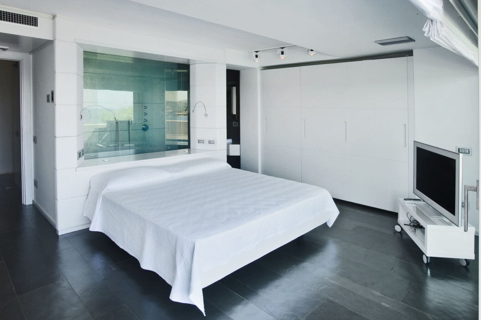 Modern minimalist bedroom featuring a white bedspread, sleek furniture, and a glass shower enclosure. The room has gray flooring, a mounted TV, and large windows allowing natural light, creating a spacious and airy atmosphere. | Only on SopranoVillas
