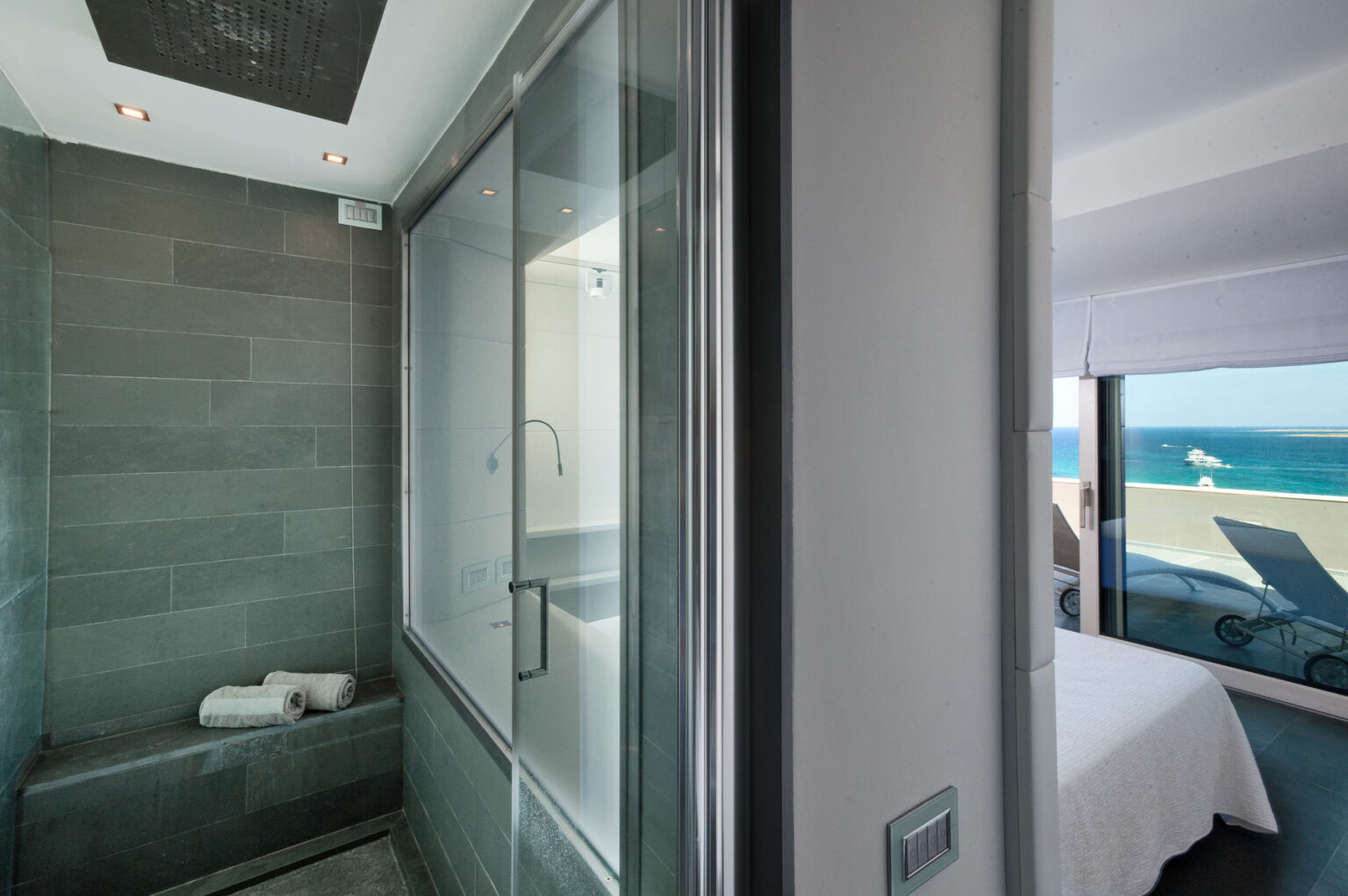 Modern bathroom featuring a spacious glass-enclosed shower with a rainfall showerhead, sleek gray tiled walls, and a comfortable seating area. The adjacent bedroom offers ocean views through large windows, enhancing the serene atmosphere. | Only on SopranoVillas