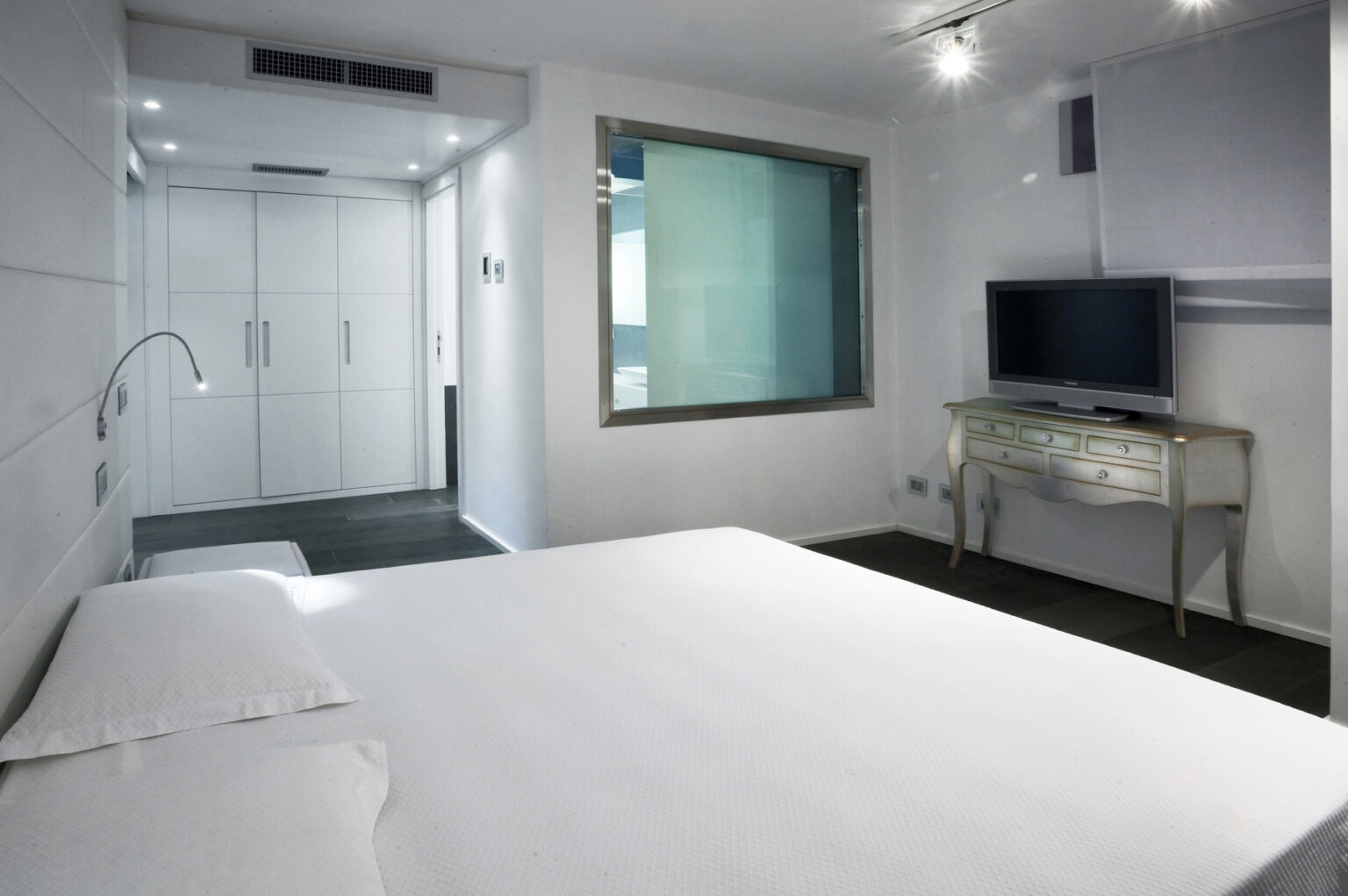 Modern hotel room featuring a spacious bed, a wall-mounted television, and a stylish desk. The room has sleek design elements with light-colored walls and a glass-enclosed shower area. | Only on SopranoVillas