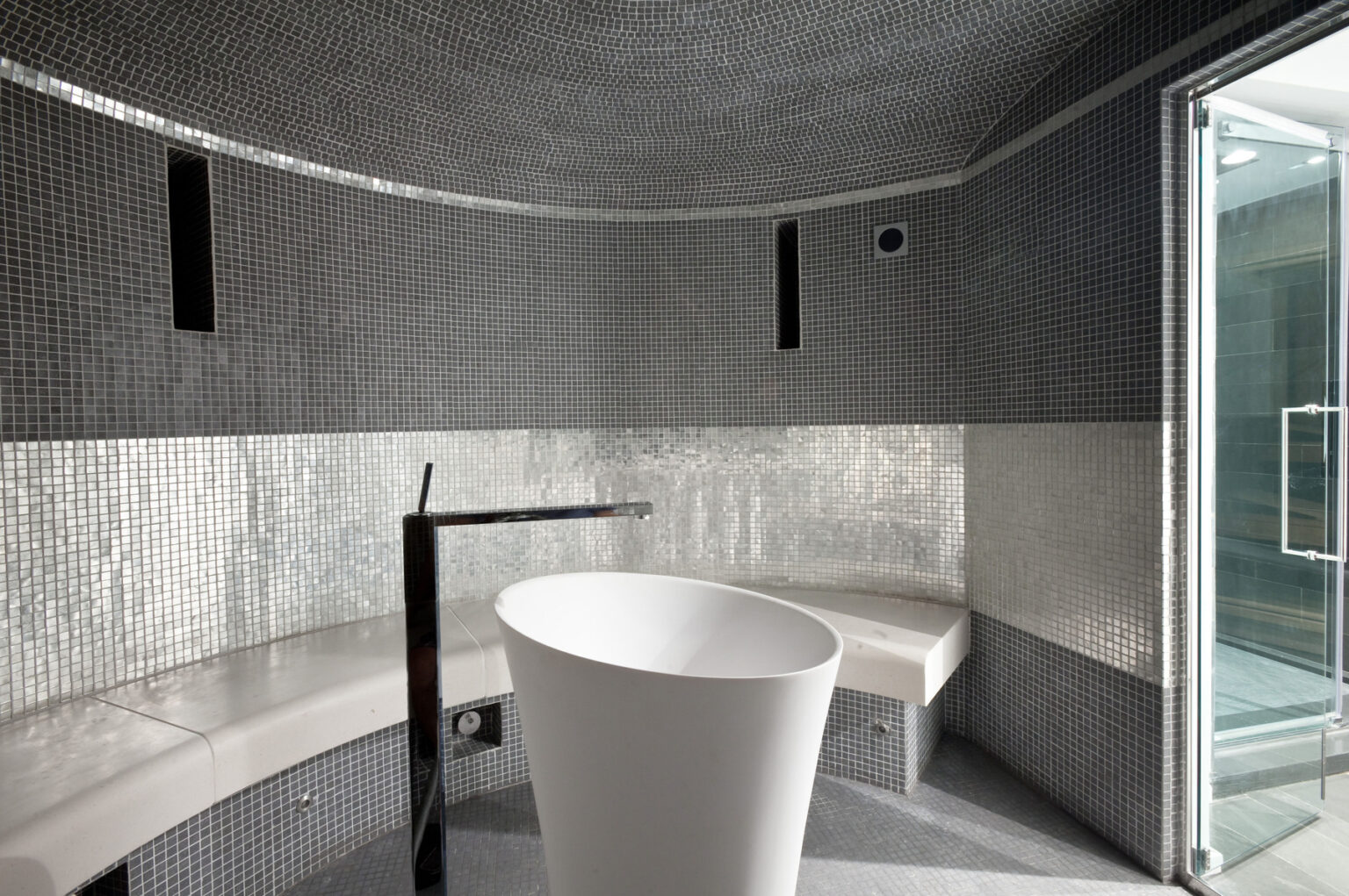 Modern steam room interior featuring sleek gray mosaic tiles, a minimalist white basin, and built-in seating. The design emphasizes relaxation and wellness, with a glass door leading to an adjoining space. | Only on SopranoVillas